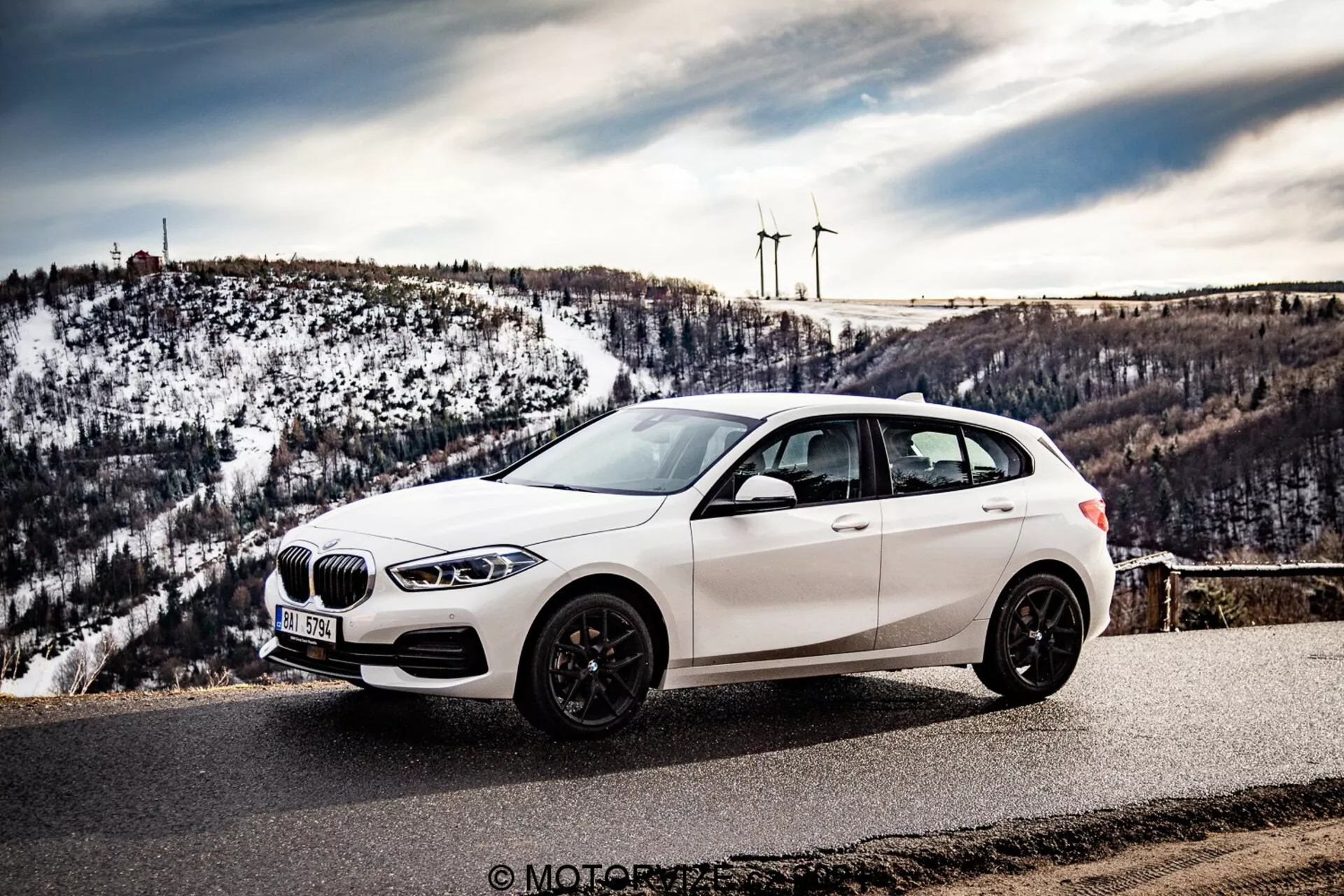 Obraz pokazuje białego hatchbacka BMW 116i z 2019 roku zaparkowanego na malowniczym punkcie widokowym lub górskiej drodze, sfotografowanego pod kątem boczno-przednim, ukazującym przedni grill, reflektory LED, przednie koło, profil boczny i spojrzenie na tylną sekcję. Perspektywa jest lekko podniesiona, co podkreśla dach samochodu, przesunięty w lewo, z tłem ośnieżonych wzgórz i odległych turbin wiatrowych. Pojazd wyposażony jest w grill w kształcie nerki, niskoprofilowe opony na wieloramiennych felgach aluminiowych, i ciemne felgi, boczne lusterka zintegrowane z kierunkowskazami oraz przyciemniane szyby. Otoczenie sugeruje jasną pogodę, co poprawia promocyjny i wizualnie atrakcyjny charakter fotografii, potencjalnie wykonanej podczas podróży lub recenzji motoryzacyjnej.