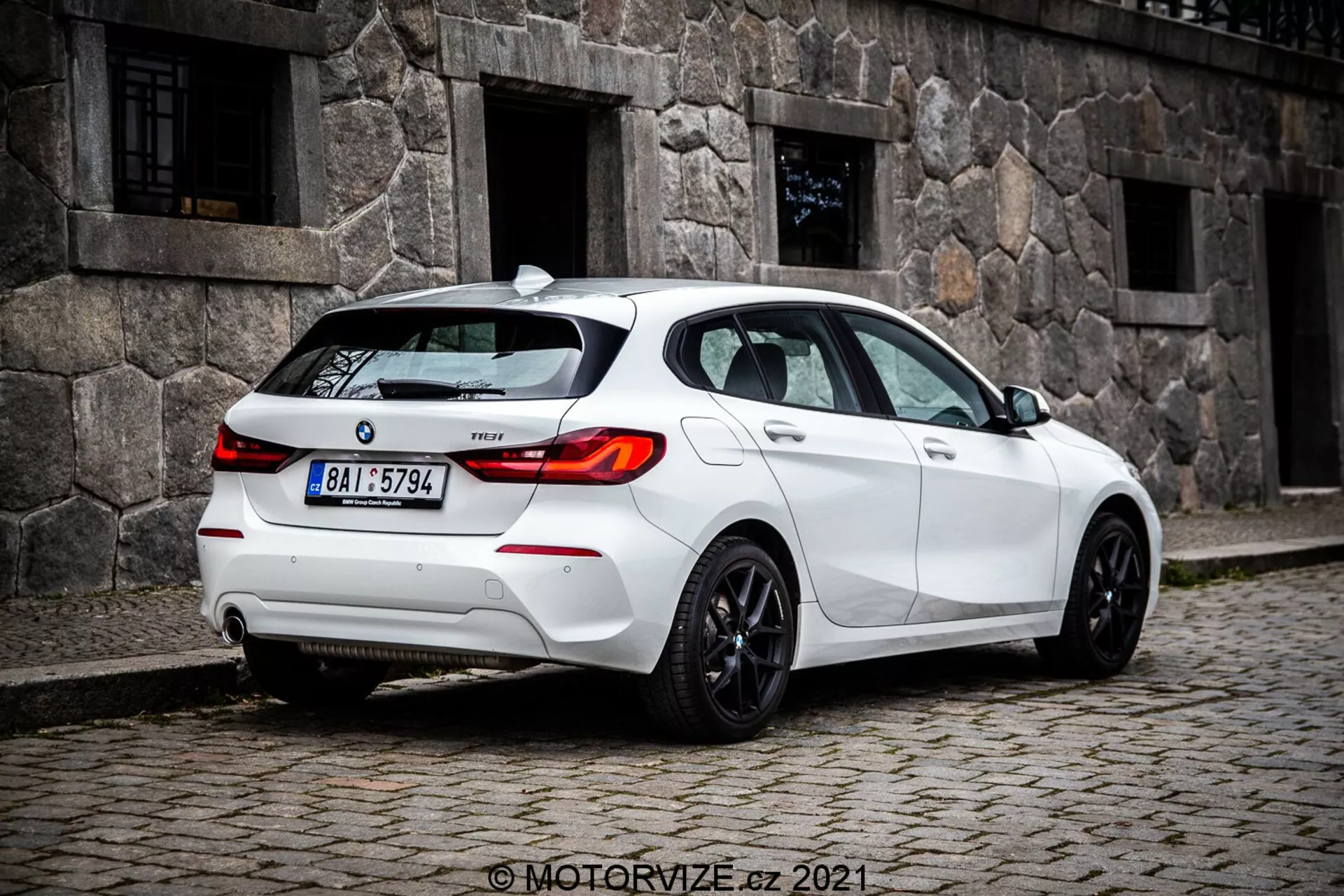 Obraz średniodystansowy na poziomie gruntu białego hatchbacka BMW 1er-serie 116i 2019, uchwycony pod kątem tylnejsko-trojkoglownej, wyraźnie pokazujący tylne światła z ledowym wzorem, tylną szybą i sportowymi felgami. Widoczne cechy obejmują czujniki w tylnym zderzaku, prawdopodobnie wspomaganie parkowania, małą antenę na dachu, prawdopodobnie dla GPS lub radia satelitarnego, odznakę modelu na bagażniku, podwójny układ wydechowy sugerujący wydajność i przyciemniane szyby dla prywatności lub komfortu.