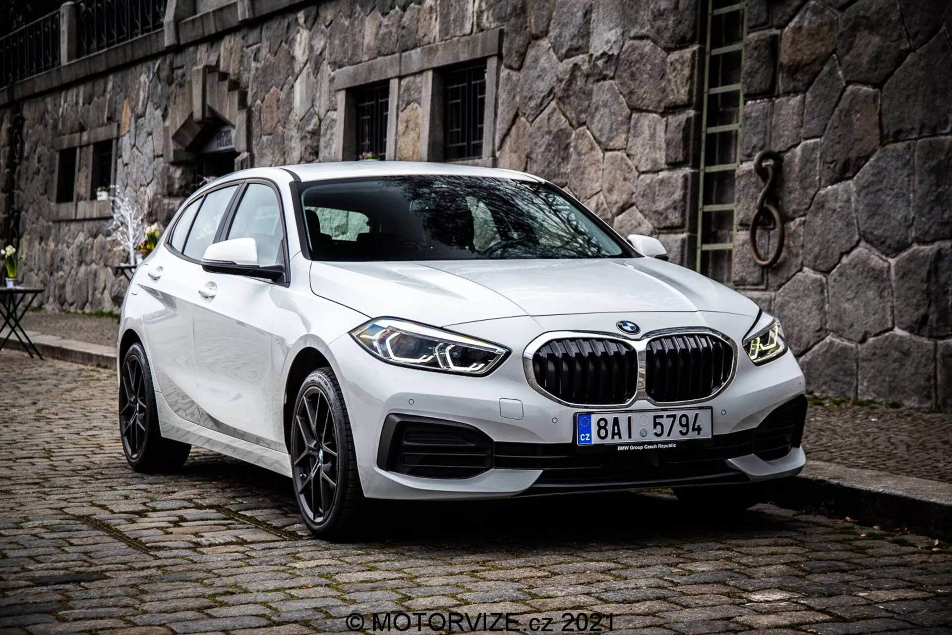 Obraz przedstawia BMW Serii 1 z 2019 roku, konkretnie BMW 116i, w białym kolorze. Zdjęcie wykonane z przedniego trójkątnego kąta pokazuje przód samochodu, stronę pasażera i szczegółowy design kół. Zdjęcie jest zrobione z nieco podwyższonego punktu widzenia, co sugeruje, że fotograf stał lub używał statywu. Samochód jest zaparkowany z przednimi kołami skręconymi w prawo, co daje wyraźny widok na obręcze i boczny profil opony. Cechy to widoczna przednia krata z pionowymi żaluzjami, rzeźbione linie na masce i bokach, wąskie reflektory LED oraz wloty powietrza na zderzaku, które mogą zawierać czujniki parkowania. Boczne lusterka z wbudowanymi kierunkowskazami i bezramowe okna podkreślają premium wygląd. Wieloszprychowe felgi aluminiowe sugerują wyższy poziom wyposażenia lub opcję ulepszoną. Stroma tylna szyba podkreśla profil hatchbacka. Pojazd jest zaparkowany na bruku w miejskiej lub historycznej scenerii z kamiennym budynkiem i małym stolikiem w stylu bistro w tle. Obraz jest ostry i szczegółowy, uchwycony w naturalnym, pochmurnym świetle dziennym.