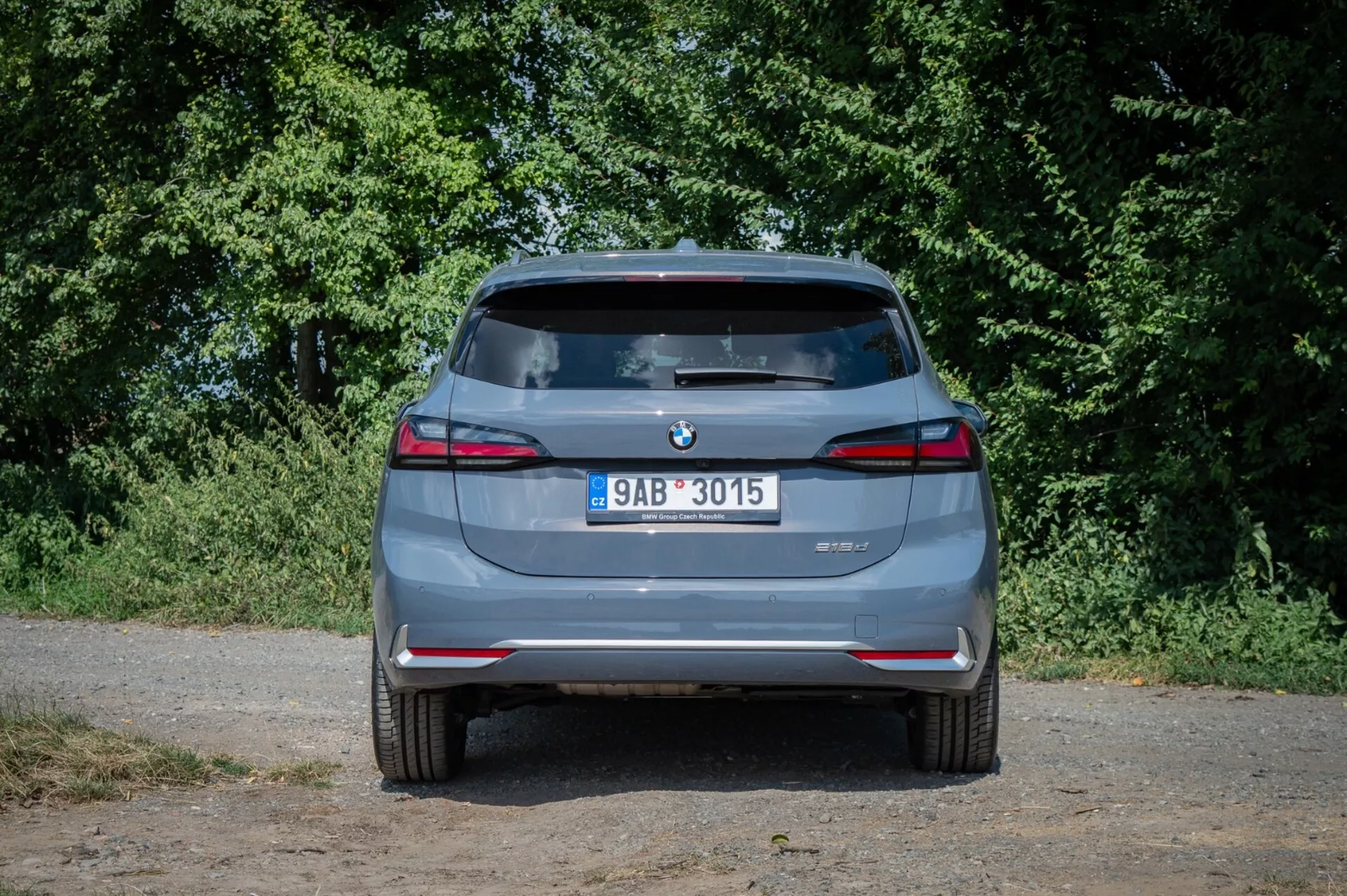 Tylny widok BMW serii 2 Active Tourer 2022, podkreślający podwójne końcówki wydechu, czujniki parkowania z tyłu i tylne światła LED w naturalnym oświetleniu z zielonym tłem podmiejskim.