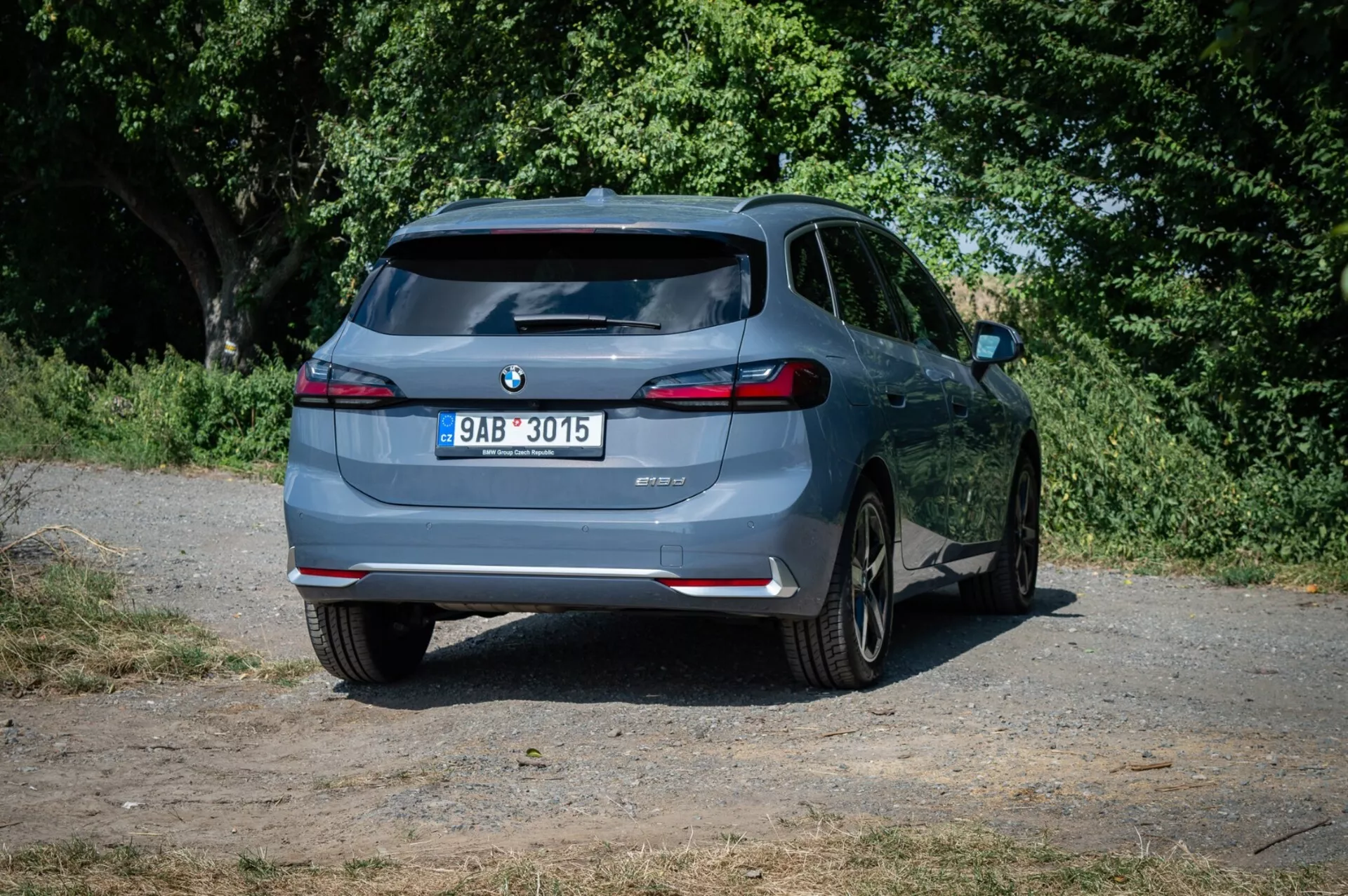 Hatchback BMW Seria 2 Active Tourer 2022, widziany z lekko przesuniętego kąta tylnego na wiejskiej drodze, podkreślający jego tylny design, tylne światła LED, podwójne wydechy i technologię wspomagania parkowania na tle natury