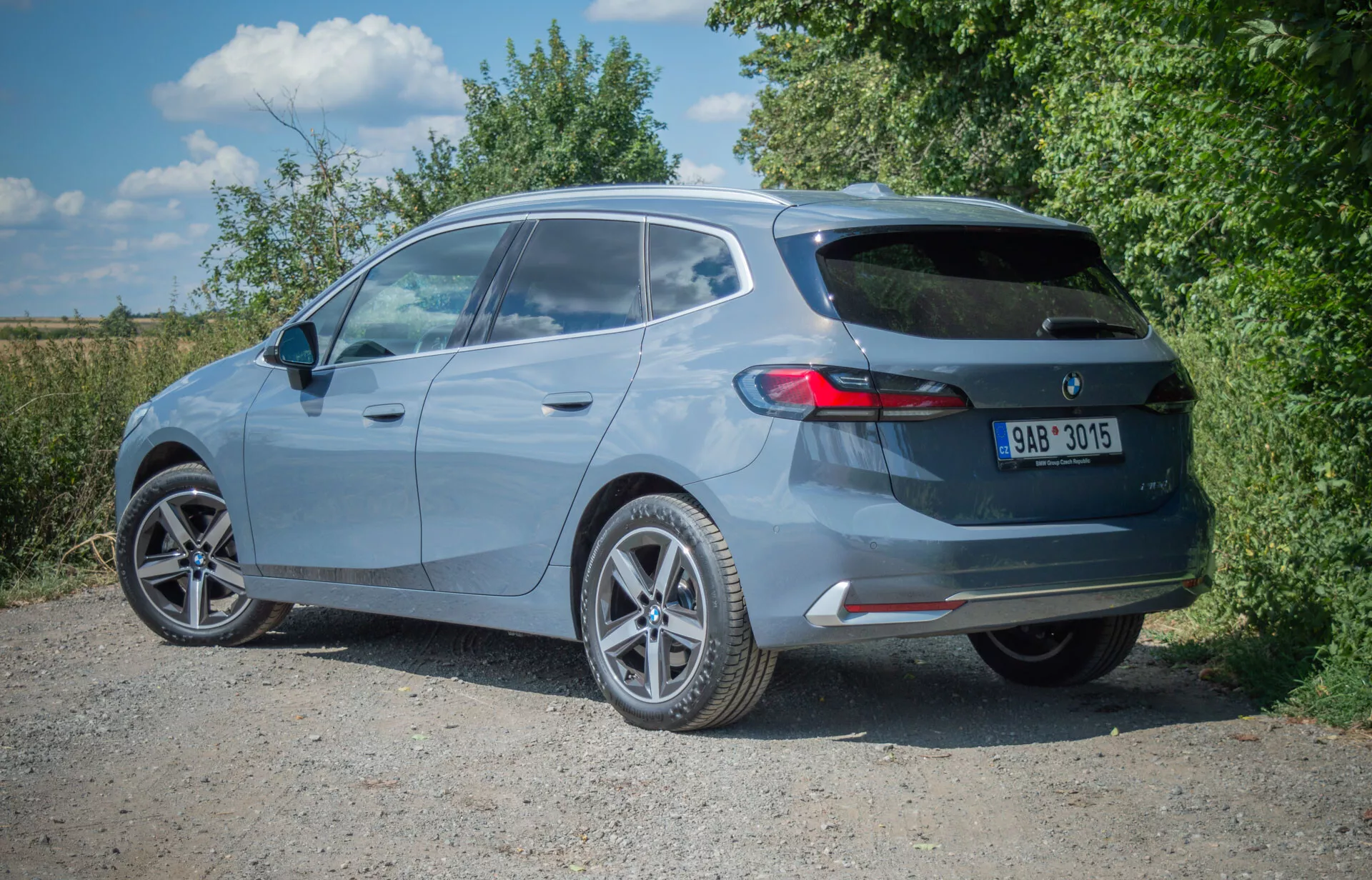 BMW Serii 2 Active Tourer hatchback 2022 w metalicznym lakierze uchwycony z perspektywy trzech czwartych tyłu, pokazujący koła ze stopu lekkiego, spoiler dachowy, tylną wycieraczkę, trzecie światło stop, poziome tylne światła, pojedynczy wydech i zderzak tylny z zintegrowanymi odblaskami, zaparkowany na nieutwardzonej nawierzchni.