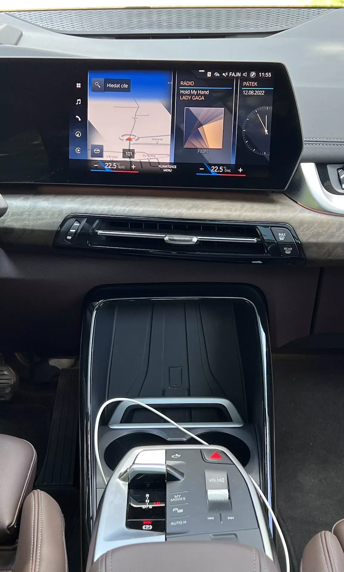 Widok wnętrza BMW Serii 2 Active Gran Tourer 2022 pokazujący centralną konsolę, szerokoekranowy dotykowy wyświetlacz infotainment z nawigacją i odtwarzaniem mediów, elegancką krateczkę wentylacyjną z drewnianym wykończeniem, przyciski do kontroli klimatyzacji, interfejs dotykowy, elektroniczny wybór biegów i premium skórzane siedzenia