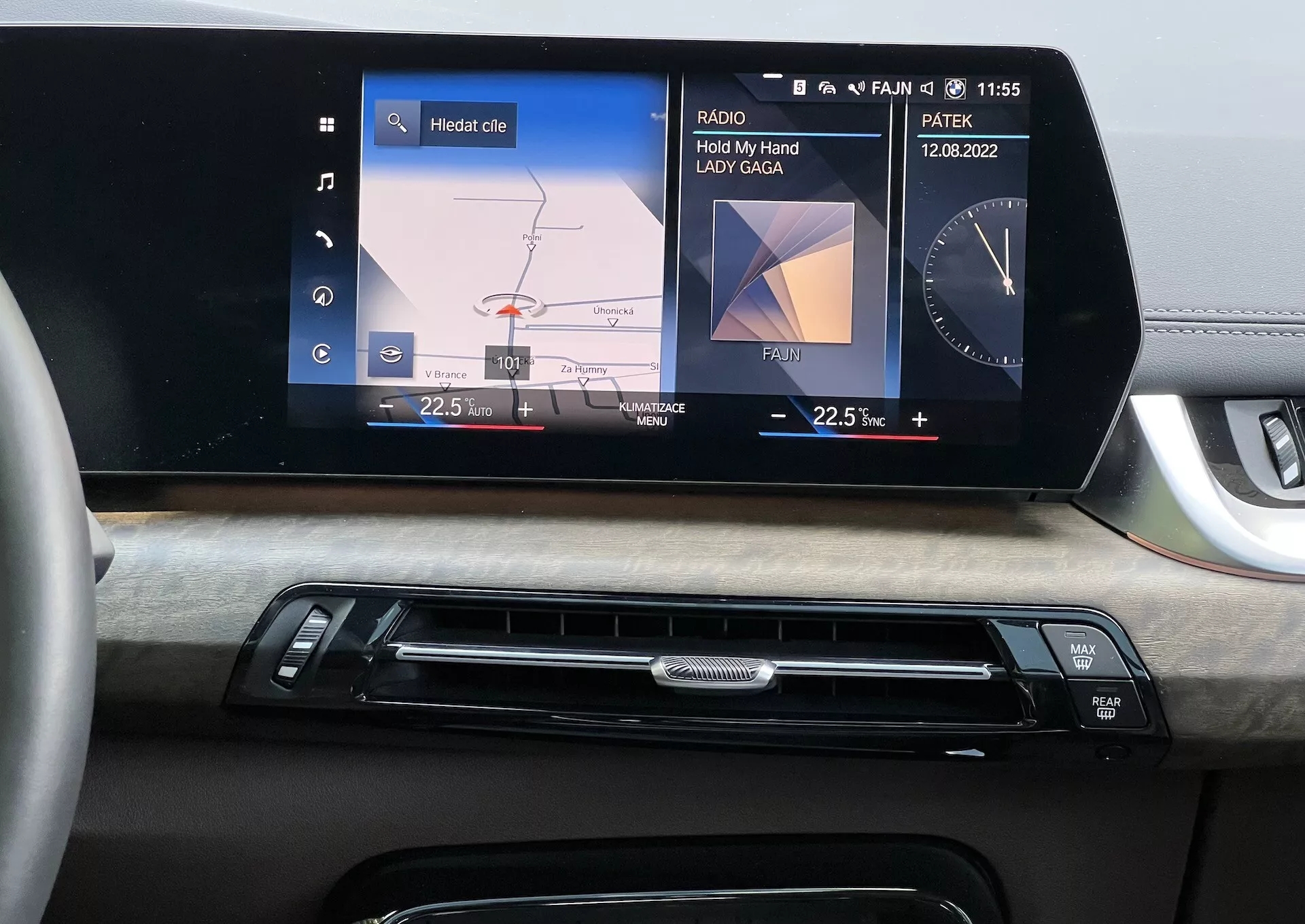Wnętrze BMW Serii 2 Active Tourer 2022 skupiające się na desce rozdzielczej z centralnym systemem infotainment pokazującym graficzne menu auta dla regulacji świateł i wyświetlacz mediów ‘Hold My Hand’ w wykonaniu Lady Gaga, widoczność daty i godziny, uzupełnione o wykończenie z drewna oraz przyciski kontroli klimatyzacji