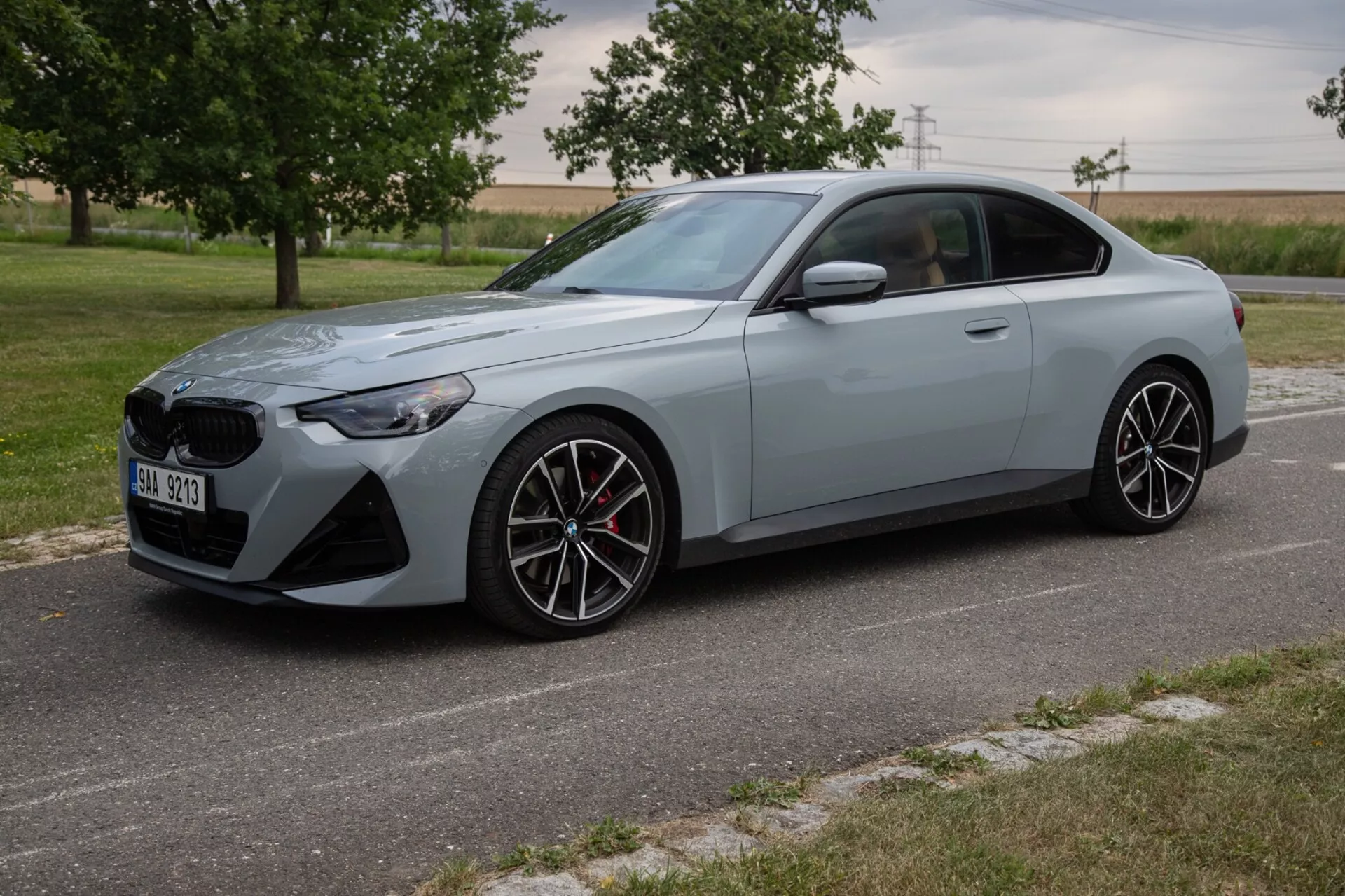 Obraz przedstawia BMW Serii 2 Coupe z 2022 roku z kąta trzech czwartych przedniego, ukazując elegancki i sportowy design z dużym przednim grillem i kątowymi reflektorami LED. Sportowe coupe, pomalowane na jasnoszary mat, wyposażone jest w wieloramienne felgi aluminiowe, sportowy układ hamulcowy z czerwonymi zaciskami oraz rzeźbione linie karoserii dla aerodynamicznego profilu. Samochód zaparkowany jest na brukowanej drodze z naturalnym krajobrazem pola w tle, w warunkach dziennego światła.