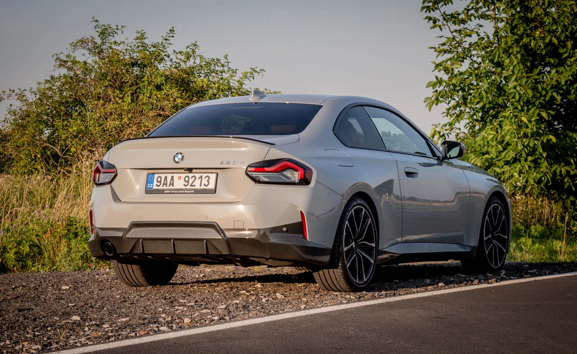 Widok tylnej części sportowego samochodu BMW 2er-reihe 2022 (2-seria coupe) uchwyconego z niskiego kąta. Samochód, zaparkowany na poboczu drogi w wiejskim otoczeniu z naturalną roślinnością, jest widoczny przy miękkim oświetleniu. Charakterystyczne znaczki identyfikują markę i model, prezentując tył, w tym pokrywę bagażnika, tylny zderzak i owinięte wokół tylne światła. Podwójne rurki wydechowe sugerują mocny silnik. Mały, zintegrowany tylny spojler i duże felgi aluminiowe z cienkimi szprychami wskazują na aerodynamiczny styl i wydajność. Metalizowany lakier samochodu i łagodnie opadająca linia dachu podkreślają jego elegancki wygląd. Widoczne nowoczesne funkcje mogą obejmować tylne światła LED, czujniki parkowania i kamerę cofania.