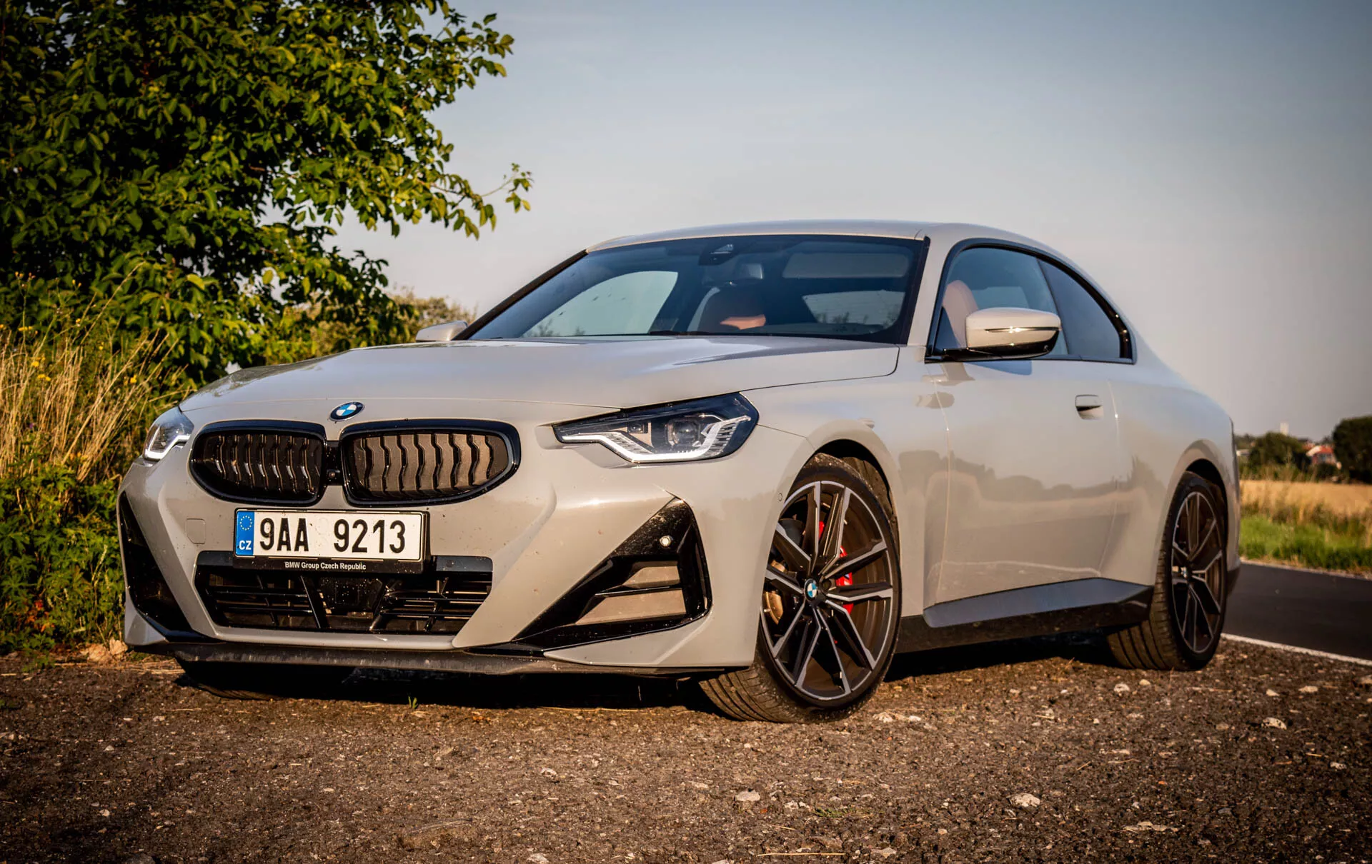Obraz nowoczesnego sportowego coupe BMW serii 2 z roku 2022. Samochód jest zaparkowany na poboczu drogi w ciągu dnia, widziany pod ukośnym kątem z lewej strony przodu, podkreślając stronę kierowcy. Cechy obejmują elegancki, aerodynamiczny design, duże, wieloramienne felgi aluminiowe z ciemnym wykończeniem, kontrastujące zaciski hamulcowe, charakterystyczny grill otoczony wąskimi, agresywnymi reflektorami LED, przednią kamerę nad tablicą rejestracyjną, duże wloty powietrza w przednim zderzaku, lekki metaliczny lakier, lusterka boczne z zintegrowanymi kierunkowskazami oraz częściowy widok na panoramiczny dach. Samochód jest ustawiony blisko aparatu z wiejskim lub podmiejskim tłem, ciepłym oświetleniem i cieniami wskazującymi na niskie słońce rano lub późnym popołudniem.