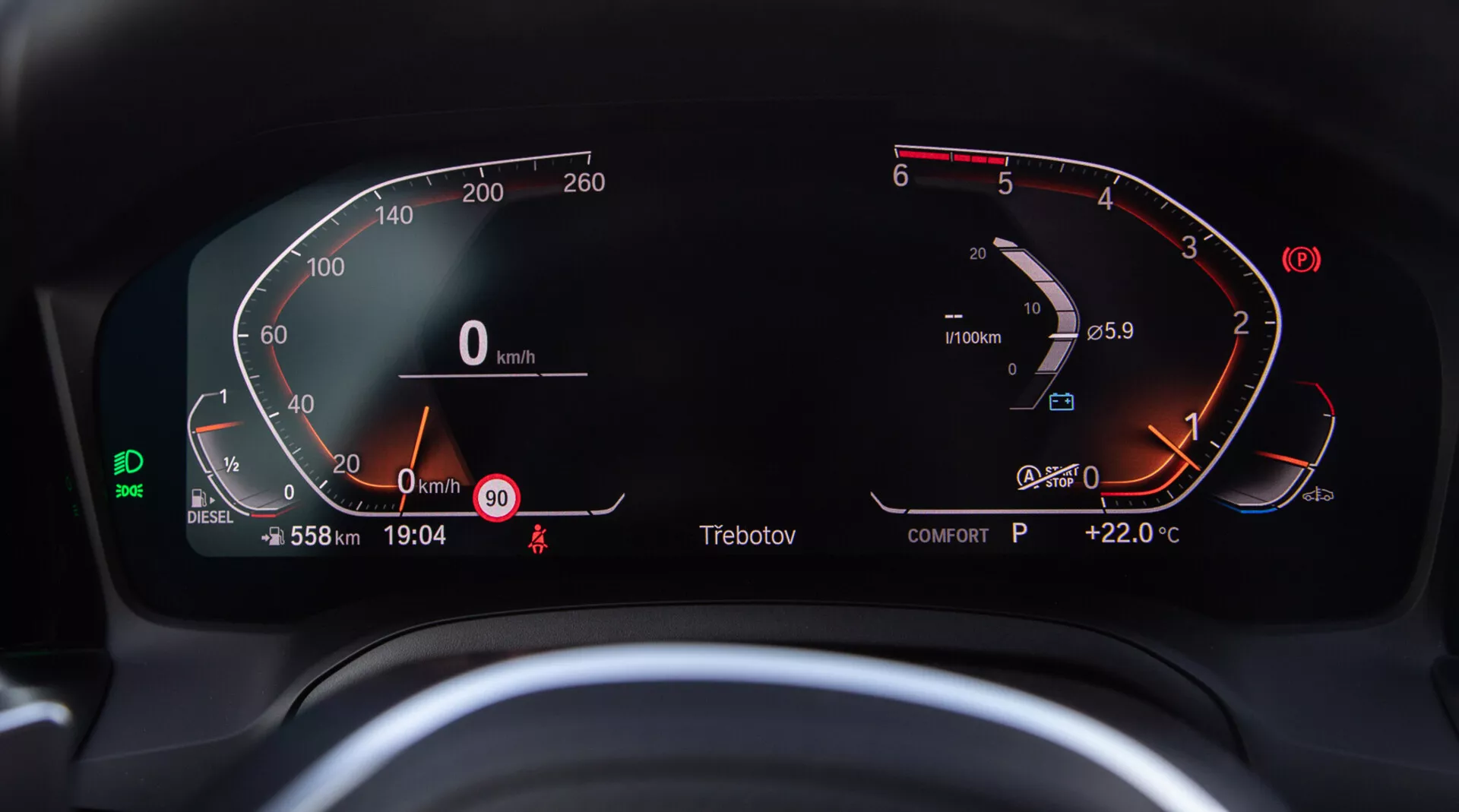 Zbliżenie na cyfrowy zestaw przyrządów w BMW 2 Series Coupe 2022, widziany z siedzenia kierowcy. Półokrągły cyfrowy prędkościomierz po lewej stronie waha się od 0 do 260 km/h, pokazując, że pojazd jest nieruchomy. Obrotomierz po prawej stronie pokazuje silnik na biegu jałowym tuż powyżej 0 RPM. Zintegrowany z obrotomierzem jest prawie pełny cyfrowy wskaźnik poziomu paliwa oraz wskaźnik temperatury silnika wskazujący na normalny zakres. Centralny wyświetlacz cyfrowy pokazuje licznik przebiegu 558 km, prędkość 0 km/h, ostrzeżenie o prędkości przy 90 km/h, aktualny czas 19:04, wskaźnik biegu 'P' na Park i temperaturę zewnętrzną +22,0°C. Widać również zieloną ikonę asystenta pasa ruchu, wyświetlacz trybu 'COMFORT', wskaźnik paliwa diesel i przypomnienie o zapięciu pasów bezpieczeństwa.