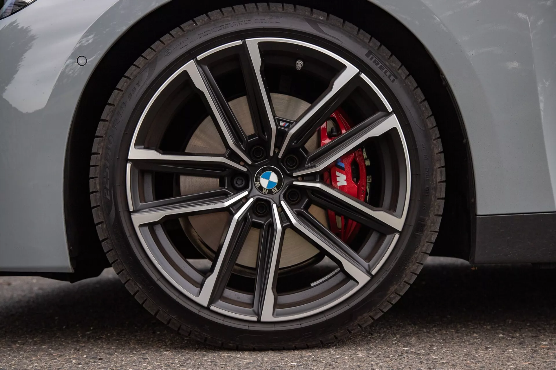 Zbliżenie koła BMW Serii 2 Coupe z wieloramienną obręczą z polerowanymi i ciemniejszymi metalicznymi wykończeniami, logo BMW na kołpaku, opona o niskim profilu oraz czerwony zacisk hamulcowy wysokiej wydajności. Jasna karoseria na asfaltowej nawierzchni podkreśla luksusowe i sportowe cechy.