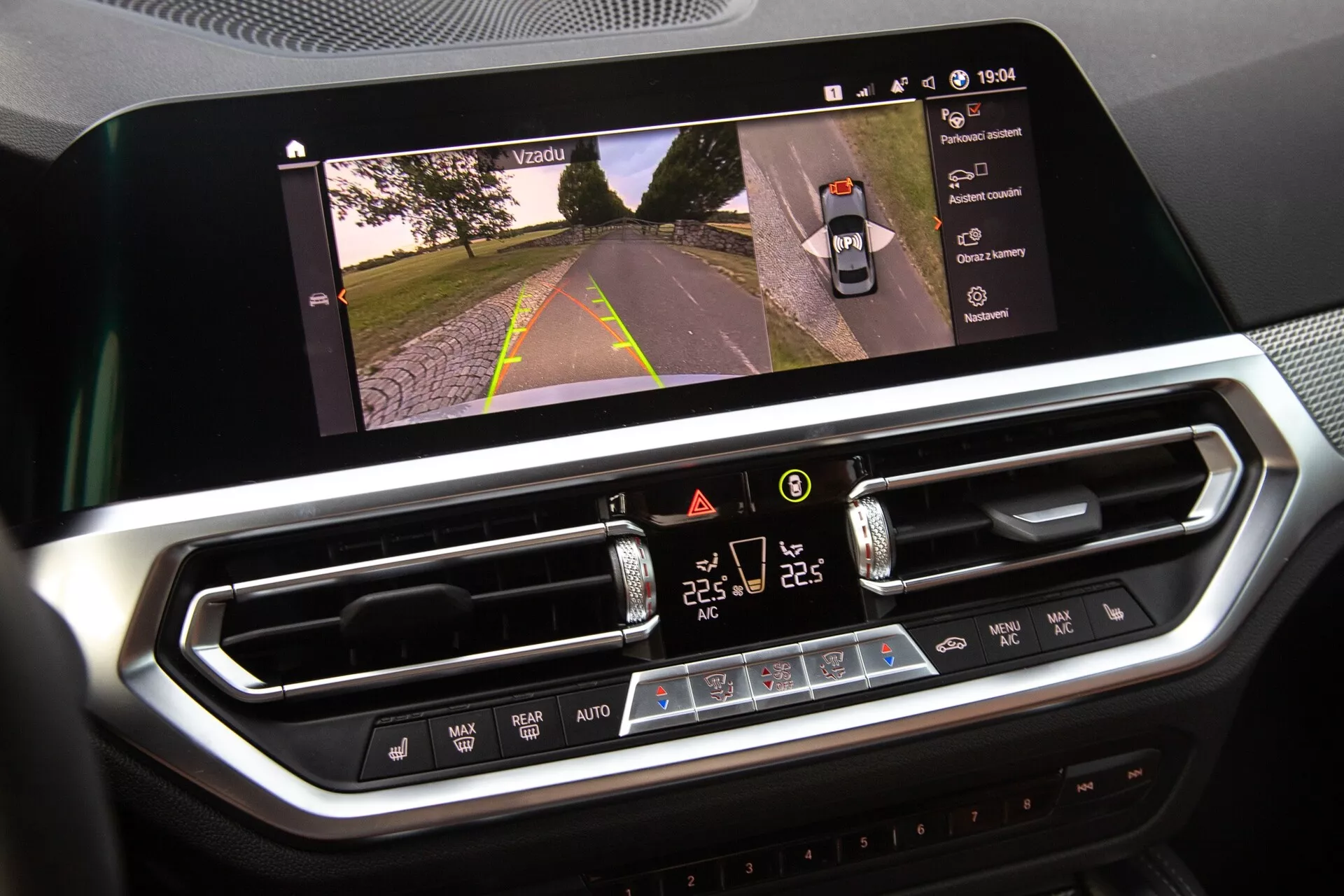 Zbliżenie wnętrza BMW 2er-Reihe 2022, ukazujące wyświetlacz infotainment oraz system kontrolowania klimatu. Duży, poziomy ekran dotykowy wyświetla widok drogi, sugerując aktywację kamery, podczas gdy cyfrowy interfejs sterowania klimatem poniżej posiada ustawienia temperatury w dwóch strefach, prędkości wentylatora i przyciski fizyczne. Nowoczesny design z chromowanymi wykończeniami sugeruje materiały premium.