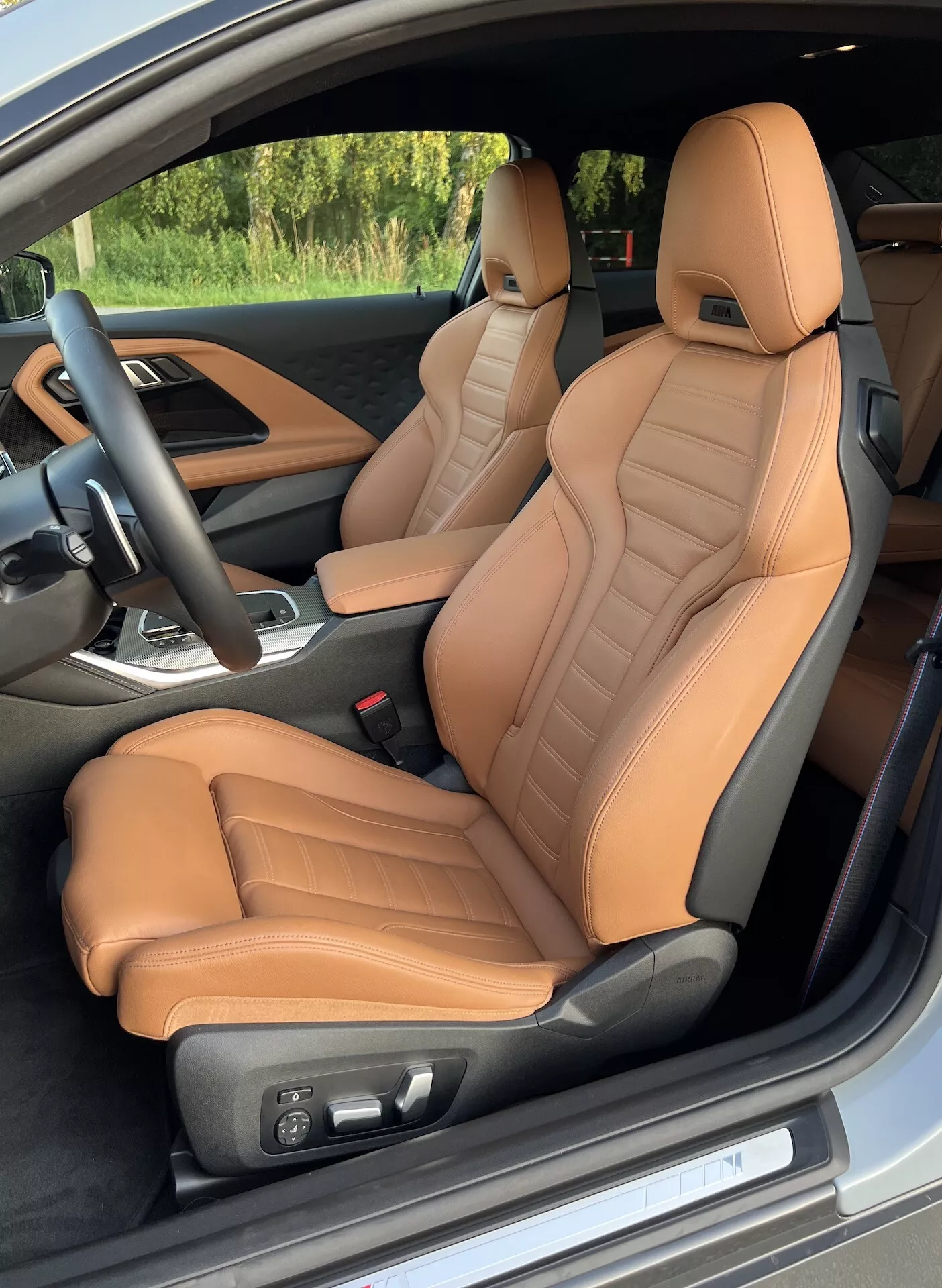 Widok wnętrza BMW Serii 2 Coupe z 2022 roku od strony drzwi kierowcy, podkreślający przednie siedzenia obite skórą w kolorze jasnobrązowym z wykończeniem pikowanym i ergonomicznym wsparciem. Elektryczna regulacja siedzeń, czarna kierownica z zamontowanymi przyciskami oraz częściowy widok centralnej konsoli z dźwignią zmiany biegów sugerują luksusowe i sportowe cechy.