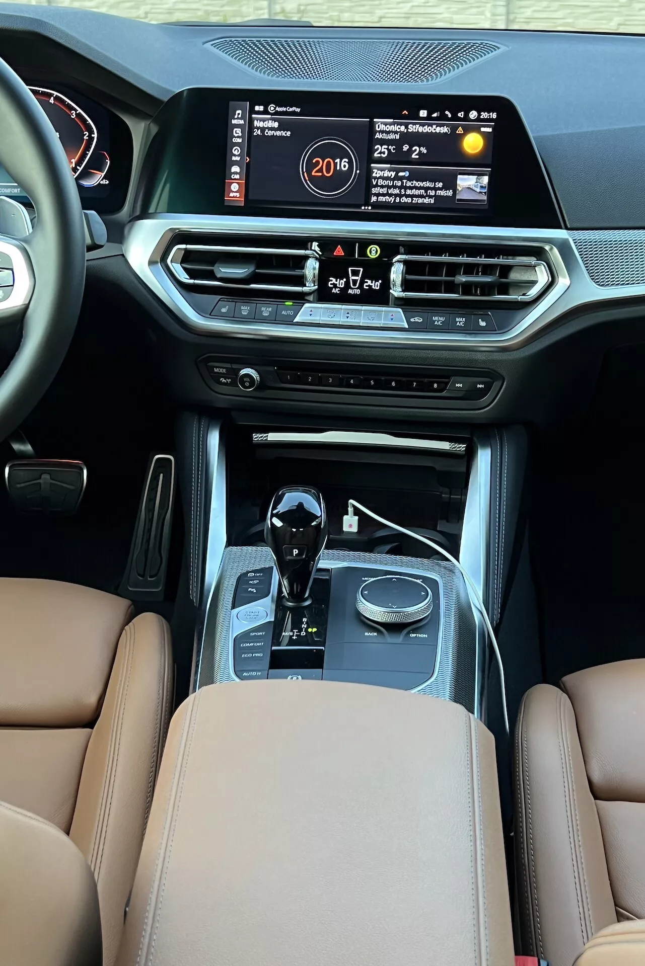 Widok wnętrza BMW Serii 2 2022 z tylnej części pasażerskiej, podkreślający środkową konsolę z dużym ekranem infotainmentu wyświetlającym czas, temperaturę i system multimedialny. Funkcje obejmują wyloty wentylacyjne klimatyzacji, przyciski sterowania klimatyzacją, przycisk start/stop, dźwignię zmiany biegów automatycznych z chromowanymi akcentami, selektory trybów jazdy, brązowe skórzane siedzenia z luksusową tapicerką i szwami, wskazujące na luksus i zaawansowaną technologię samochodową.