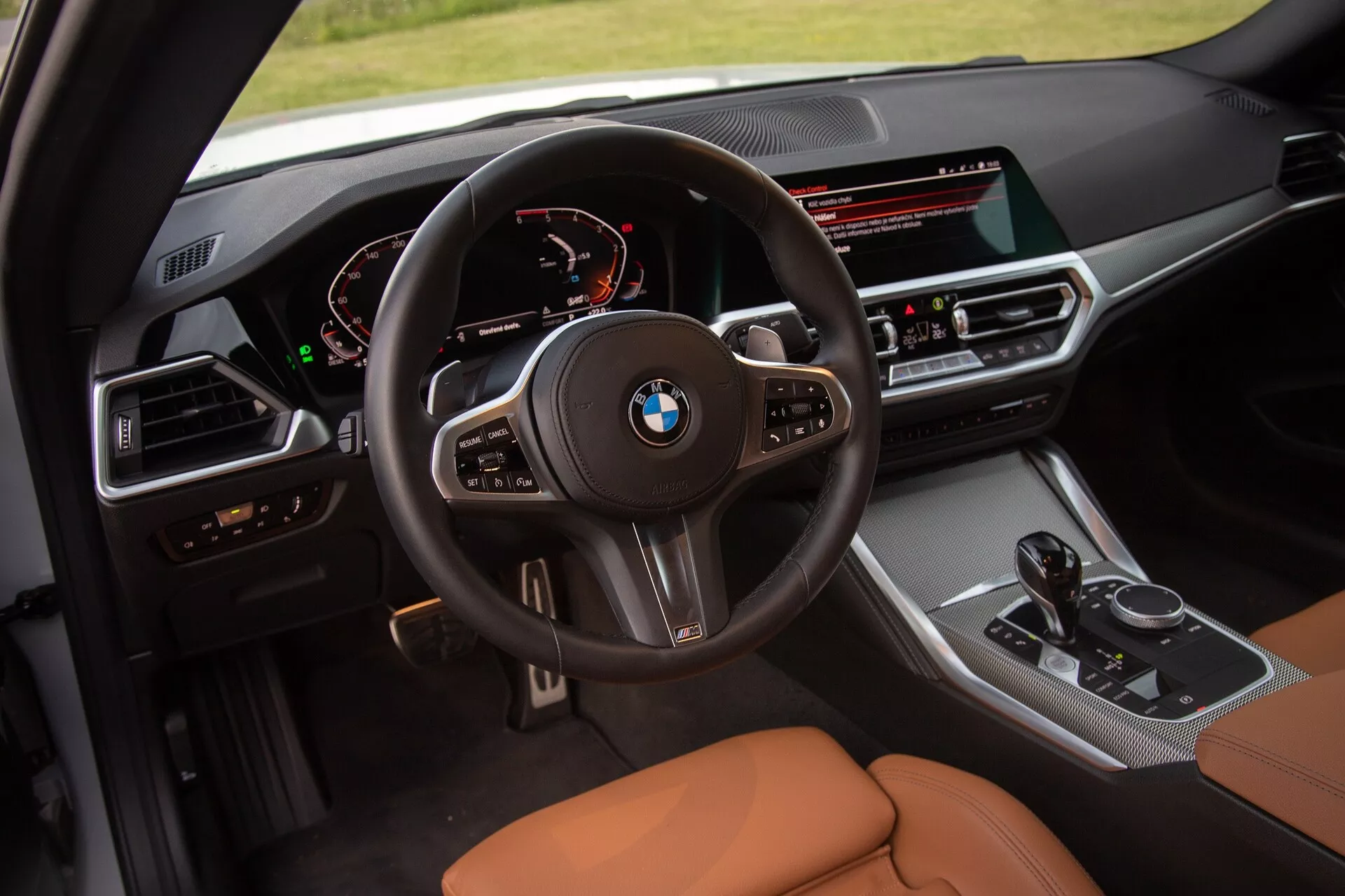 Zdjęcie wnętrza BMW Serii 2 Coupé 2022 skupiające się na kokpicie kierowcy. Zdjęcie z podwyższonej perspektywy ukazuje szczegółowy widok wielofunkcyjnej kierownicy z logo BMW, cyfrowego zestawu wskaźników wyświetlającego prędkościomierz i obrotomierz, oraz zorientowanej na kierowcę konsoli środkowej z szerokim ekranem infotainment, przyciskami sterowania klimatyzacją, dźwignią zmiany biegów oraz pokrętłem sterowania iDrive. Siedzenia są wyściełane karmelem lub brązową skórą, podkreślając luksus i komfort.