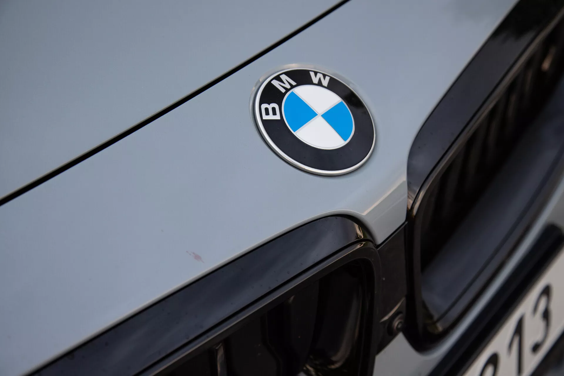 Zbliżenie przedniej części BMW 2-Series Coupe 2022, z wyraźnie umiejscowionym na środku czarnym, niebieskim i białym emblematem BMW na masce. Obraz uchwyca górną krawędź ikonicznego grilla nerkowego w tle, z metalicznym, srebrnym lub jasno szarym kolorem nadwozia odbijającym światło otoczenia. Ostre ustawienie ostrości na emblemacie podkreśla luksusową markę i dobrze utrzymaną powierzchnię pojazdu.