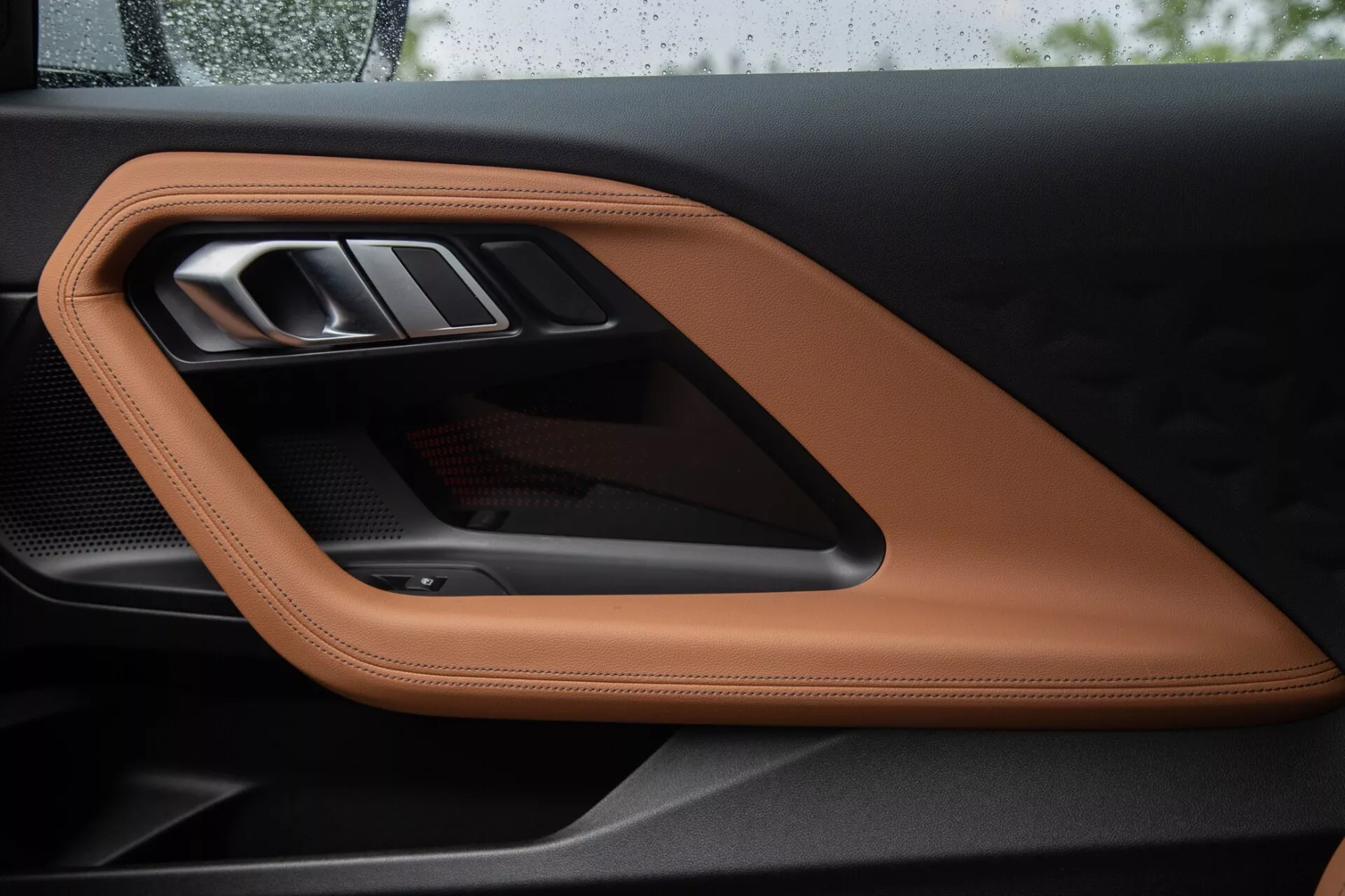 Zbliżenie na panel drzwi wewnętrznych BMW 2 Series Coupe 2022, ukazujący luksusową przeszywaną skórę w kolorze brązowym lub koniakowym, elegancką metalową klamkę drzwi i sportowy akcent z włókna węglowego, podkreślający wysokiej jakości materiały i wyrafinowane elementy designu.