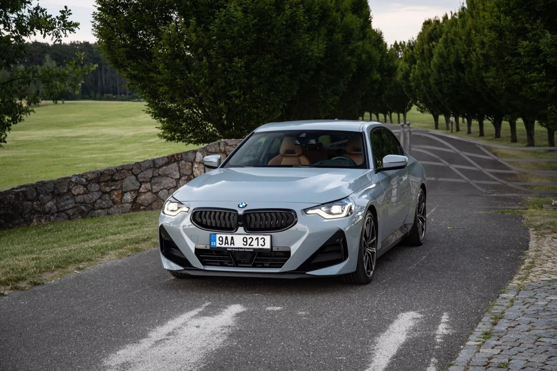Szczegółowy obraz BMW Serii 2 Coupé 2022 uchwycony z przodu pod kątem trzech czwartych, prezentujący przednie i boczne profile na spokojnej asfaltowej drodze, otoczonej przyciętymi drzewami i kamiennym murem. Charakterystyczna dla BMW nerka i eleganckie reflektory LED zdobią przednią część, a wloty powietrza w zderzaku podkreślają sportowy wygląd. Profilowane linie maski sugerują osiągi, wyraźnie zarysowane błotniki wskazują na moc, a wieloramienne felgi aluminiowe oznaczają sportową wersję. Profil boczny prezentuje linię charakteru, która daje wrażenie ruchu, lusterka boczne z wbudowanymi kierunkowskazami i zarys szyberdachu. Standardowy kąt nachylenia szyby przedniej i przezroczyste boczne okna dodają zrównoważony wygląd. Delikatne oświetlenie, prawdopodobnie wcześnie rano lub późnym popołudniem, tworzy subtelne cienie i czystą prezentację designu i koloru samochodu.