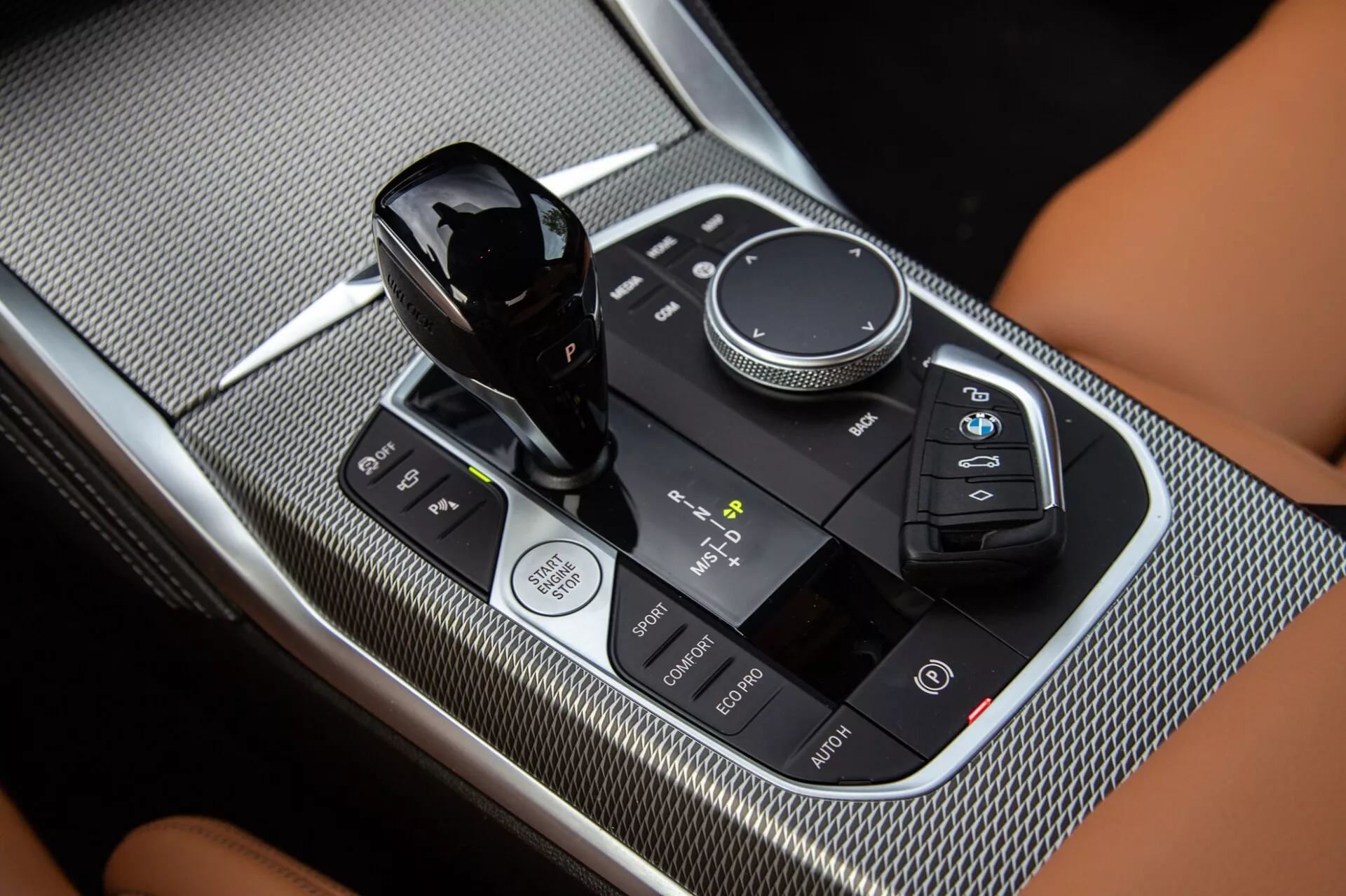 Zbliżenie konsoli centralnej w BMW 2er-Reihe 2022, uwydatniające dźwignię zmiany biegów, pokrętło sterowania multimediami i dotykowy panel sterowania z powierzchniami z połyskiem i teksturą z włókna węglowego.