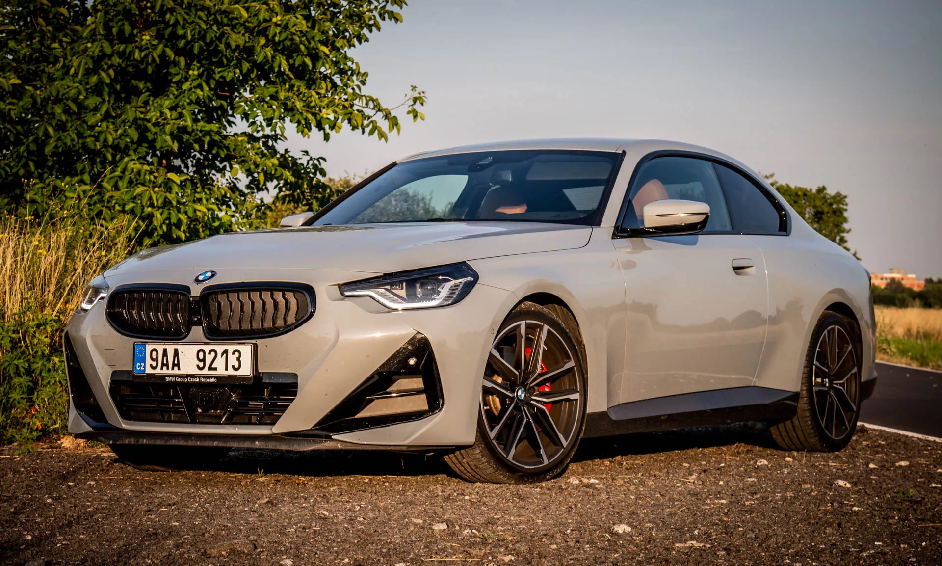 Obraz BMW 2 Series Coupe z 2022 roku. Nowoczesny luksusowy samochód jest zaparkowany pod kątem, podkreślając przednią lewą stronę podczas złotej godziny. Ujęcie skupia się na charakterystycznych elementach designu, takich jak duża nerka w osłonie chłodnicy, kanciaste reflektory LED, sportowy przedni zderzak z dużymi wlotami powietrza oraz wieloramienne czarne felgi aluminiowe z wysokowydajnymi zaciskami hamulcowymi. Aerodynamiczne lusterko boczne ma zintegrowane kierunkowskazy. Elegancka karoseria i wysokiej jakości wykończenie lakieru podkreślają kompaktowy, sportowy profil samochodu i niską linię dachu.