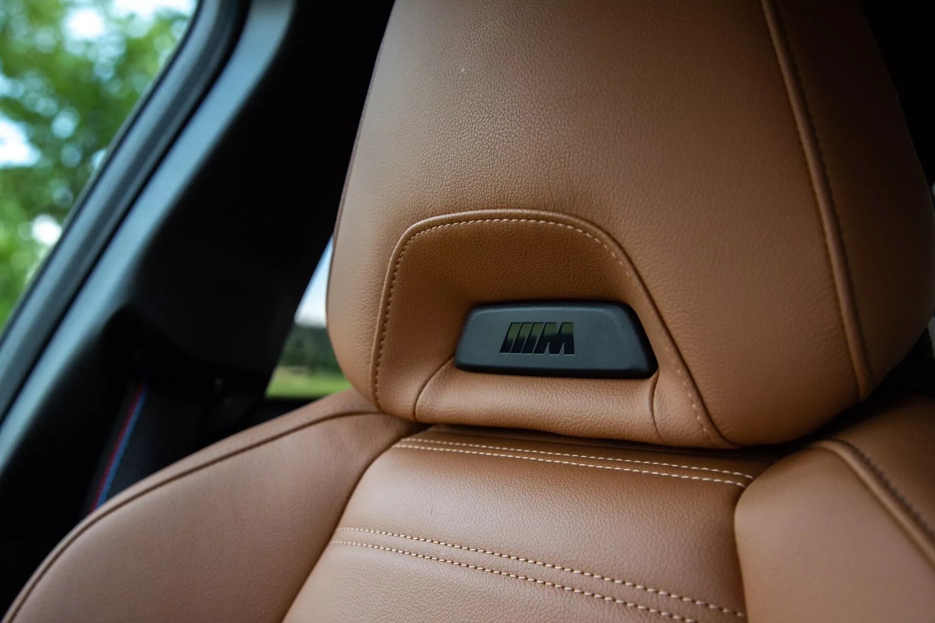 Zbliżenie górnej części brązowej skórzanej tapicerki w BMW 2 Series Coupe 2022, podkreślające precyzyjne przeszycia oraz ergonomicznie zaprojektowaną linię konturu dla komfortu i wsparcia. Na górnej części siedzenia znajduje się plakietka osadzona w wytłoczeniu, wskazująca na funkcje technologiczne, takie jak regulacja elektroniczna lub funkcje grzania. Zdjęcie wykonano z nieco niższego punktu widzenia, podkreślając luksusową fakturę skóry przy jednoczesnym rozmyciu tła, które obejmuje część okna auta.
