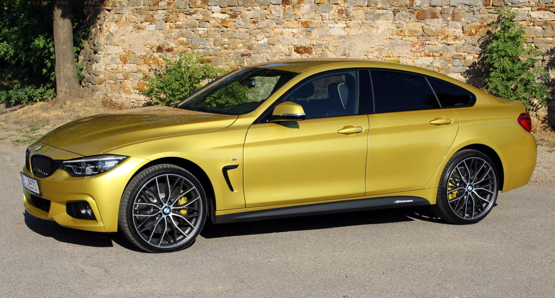 Metalicznie złote coupé BMW Serii 4 z 2013 roku zaparkowane równolegle do starego kamiennego muru, pokazujące swój elegancki, aerodynamiczny profil boczny, felgi aluminiowe i ulepszenia technologiczne z nieco podniesionego kąta