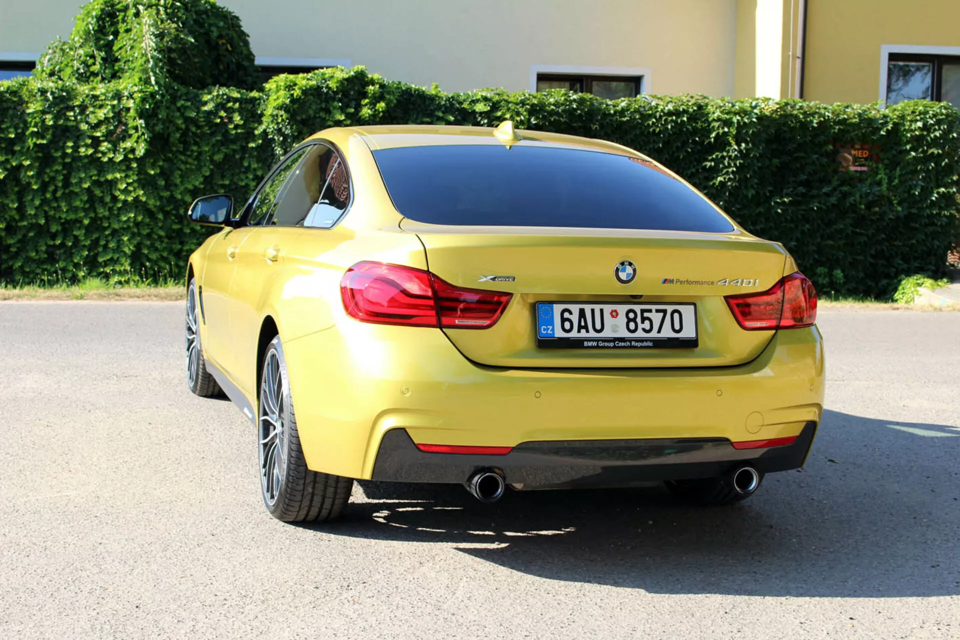 Żółty BMW Serii 4 Coupé z 2013 roku z tylnej perspektywy trzy czwarte ukazujący projekt bagażnika, tylny zderzak, część tylnej nadkoli, charakterystyczne tylne światła, podwójne rury wydechowe, tablicę rejestracyjną, odznakę BMW, oznaczenie modelu, czujniki parkowania, antenę płetwą rekina oraz felgi z lekkich stopów o wieloramiennej konstrukcji, zaparkowane w miejskim otoczeniu w słoneczny dzień.
