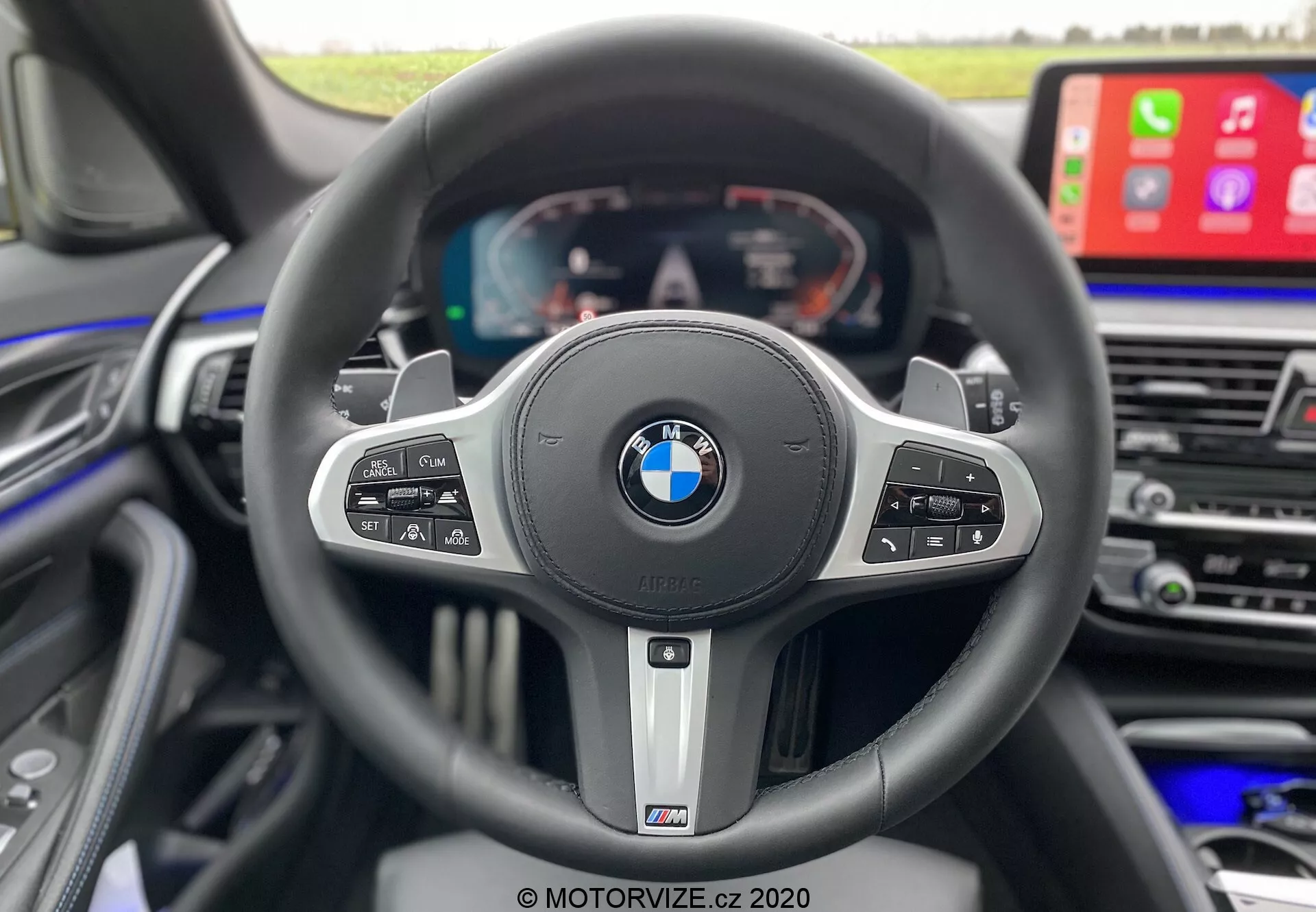 Widok wnętrza z perspektywy kierowcy wewnątrz BMW 520i Touring Steptronic 2020, wyróżniający sportową, trzyramienną kierownicę z emblematami BMW i M. Kierownica wyposażona jest w liczne przyciski sterujące do tempomatu, mediów, poleceń głosowych i funkcji telefonicznych, oraz łopatki zmiany biegów wskazujące na automatyczną skrzynię biegów z opcją manualną. Za kierownicą widać w pełni cyfrowy wyświetlacz pokazujący prędkościomierz, obrotomierz i informacje o pojeździe. Po prawej widoczna jest część centralnej konsoli z dużym ekranem infotainmentowym pokazującym Apple CarPlay, podkreślając luksus, zaawansowaną technologię i projekt skoncentrowany na kierowcy.