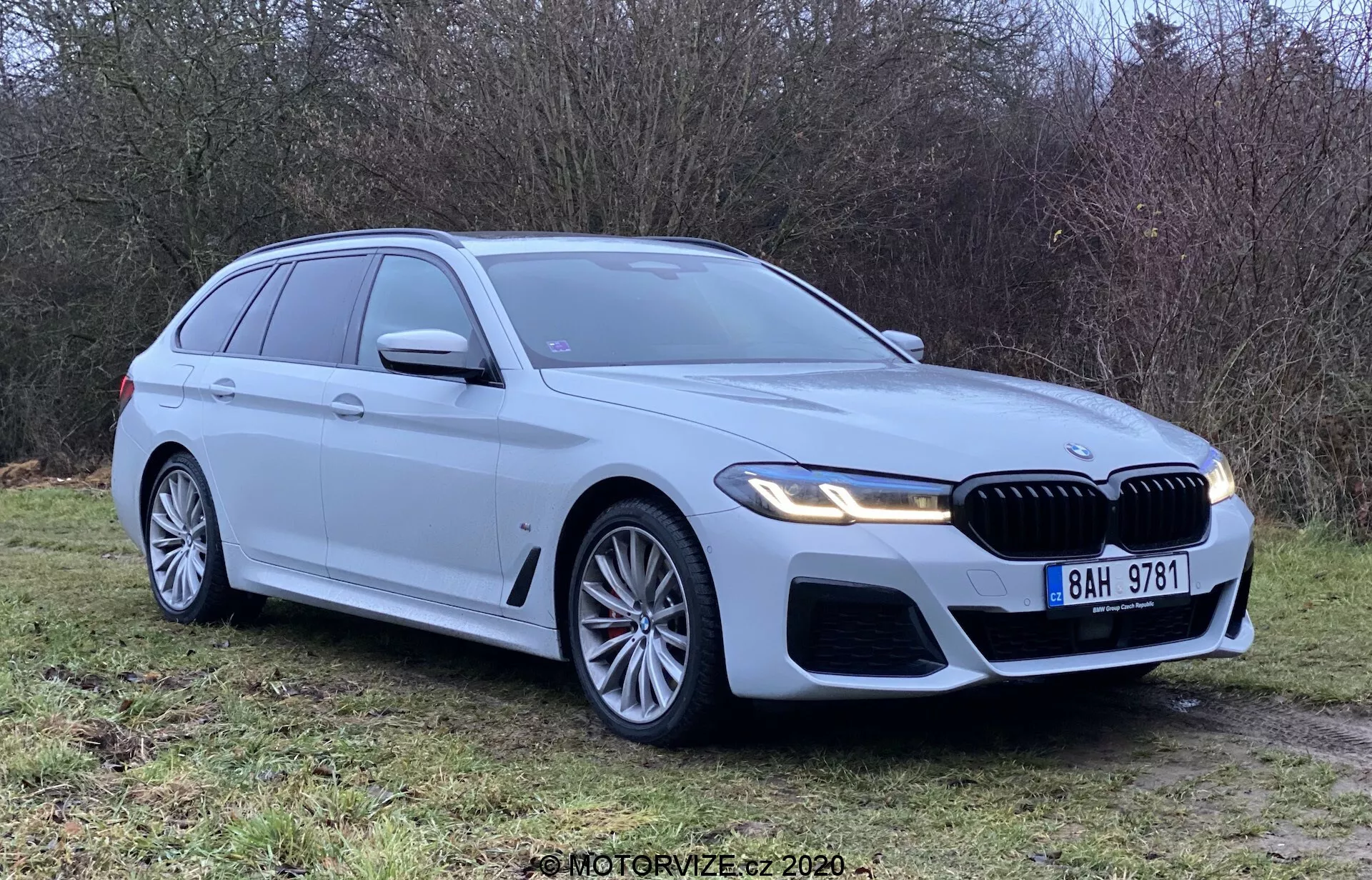 Srebrny BMW 5 Series Touring 2020 zaparkowany na trawie, widziany pod kątem trzech czwartych z przodu, pokazujący przedni grill, reflektory i aerodynamiczny design. Wyposażony w reflektory LED, duże felgi aluminiowe, grill „nerki” i czujniki parkowania. Czysty i nowy, podkreślający znak BMW, zintegrowane kierunkowskazy, relingi dachowe i antenę w kształcie płetwy rekina.