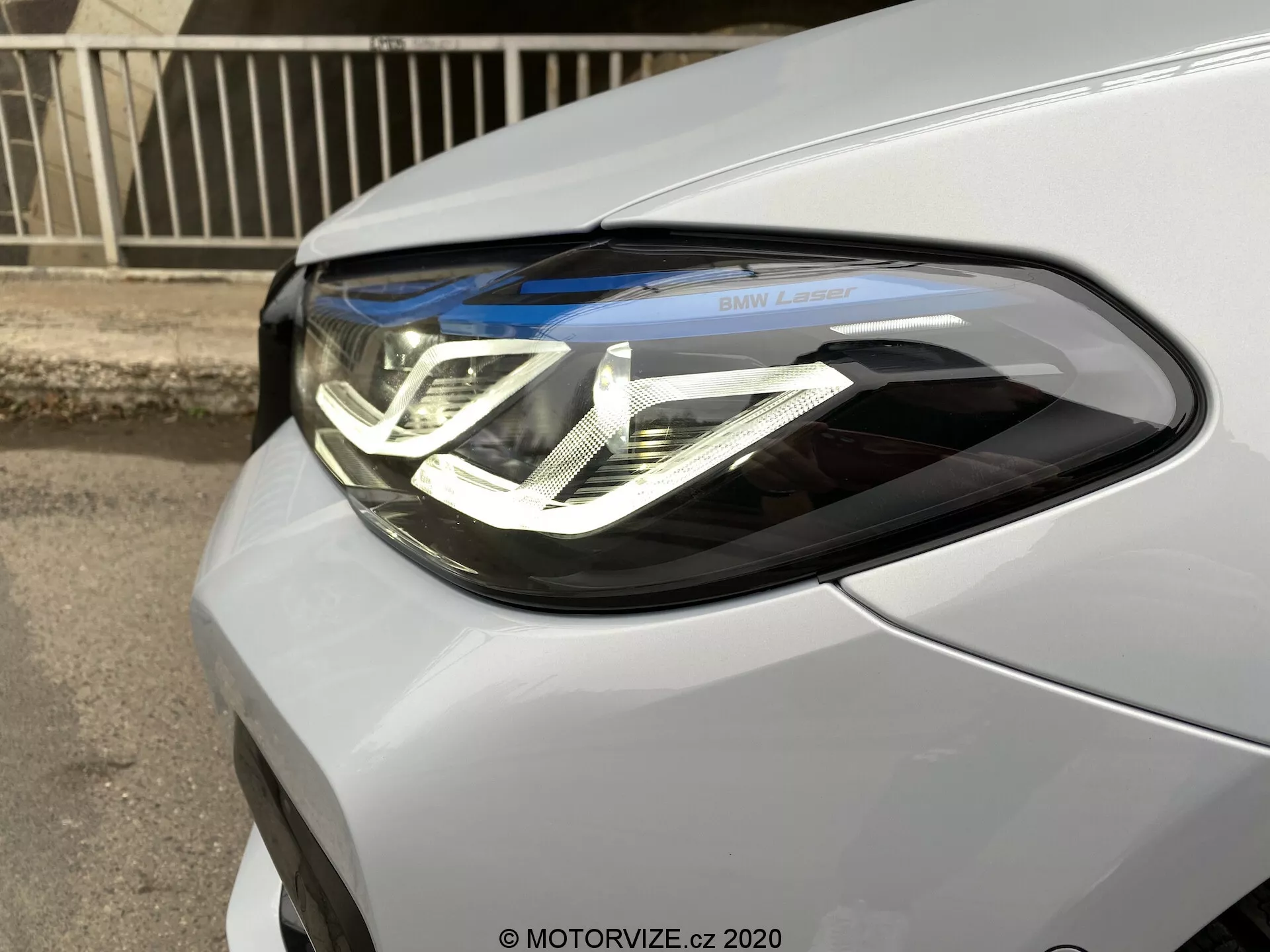 Zbliżenie reflektora po stronie kierowcy BMW serii 5 2020 (BMW 520i Touring Steptronic). Obraz uchwyca samochód pod kątem trzech czwartych z przodu, podkreślając skomplikowane światła do jazdy dziennej LED z podwójnym kształtem litery U, soczewki projektora i napis 'BMW Laser'. Jednostka reflektora bezszwowo integruje się z metalicznym lub perłowym lakierem samochodu.
