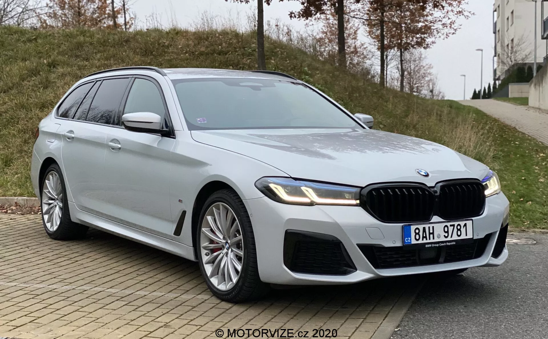 Nowoczesne, luksusowe BMW Serii 5 (BMW 520i Touring Steptronic) kombi sfotografowane z niskiego kąta z przodu z trzech czwartych. Obraz podkreśla przedni grill samochodu, eleganckie, kanciaste reflektory prawdopodobnie wyposażone w technologię LED oraz muskularne linie projektowe na masce i błotnikach. Widoczne części obejmują przedni grill, zderzak, reflektory, drzwi kierowcy z przodu, przedni błotnik, część tylnych drzwi, okna boczne, koła i lusterko boczne po stronie kierowcy. Duże, aluminiowe felgi sugerują sportową wersję modelu, a srebrny lakier jest odbijający i czysty. Samochód ma wyraźny przedni grill z pionowymi listewkami, plakietkę na przednim błotniku sugerującą wyższy poziom wykończenia oraz lusterka boczne z wbudowanymi kierunkowskazami. Widoczny jest również szyberdach. Pojazd jest zaparkowany przy miejskiej drodze z krawężnikiem i chodnikiem na pierwszym planie oraz trawiastym terenem i budynkiem w tle, w pochmurnych warunkach oświetleniowych.