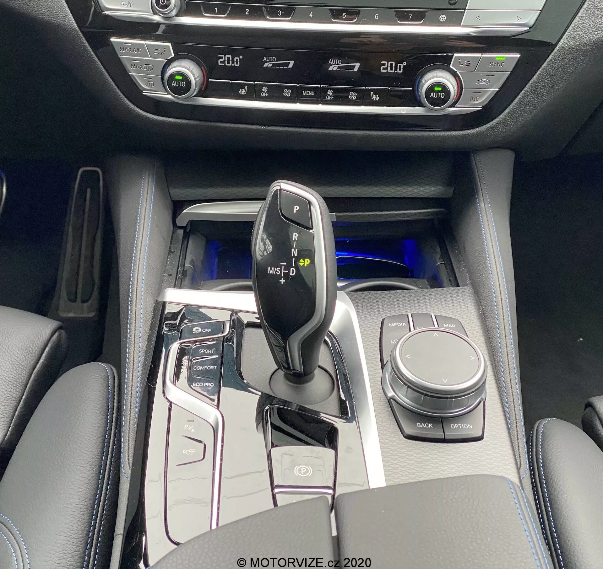 Widok z lotu ptaka na konsolę centralną BMW 520i Touring Steptronic 2020, przedstawiający błyszczący czarny drążek zmiany biegów z metalicznymi akcentami, otoczony panelem sterowania o wysokim połysku z przyciskami do systemu start/stop, trybów jazdy, czujników parkowania i więcej. Widoczny jest pokrętło iDrive, uchwyty na kubki i wyściełany podłokietnik z miejscem do przechowywania. Chłodne niebieskie oświetlenie ambientowe podkreśla luksusowy wygląd. Kontrola klimatyzacji dwustrefowej i przyciski grzania siedzeń wskazują na zaawansowane funkcje komfortu. Materiały wewnętrzne obejmują powierzchnie podobne do skóry z metalicznymi akcentami i błyszczącymi panelami.