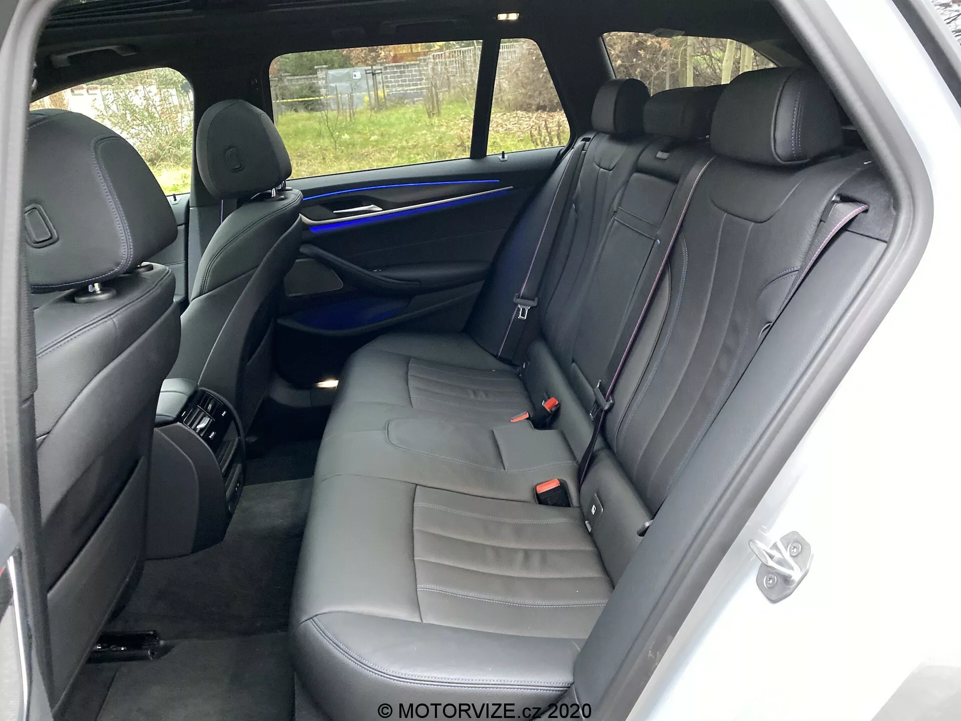 Wnętrze BMW 520i Touring Steptronic z 2020 roku skupiające się na tylnych siedzeniach z prawej strony. Punkt widzenia znajduje się blisko tylnego prawego drzwi pasażera, pokazując ławkę dla trzech osób z ciemną skórzaną tapicerką i ozdobnymi przeszyciami. Funkcje obejmują trzy zagłówki, trzy pasy bezpieczeństwa, panel drzwiowy z kratką głośnika, uchwyt i elementy sterujące, niebieski pasek oświetlenia ambientowego oraz siatkę do przechowywania na tylnej stronie przedniego siedzenia pasażera. Dobrze utrzymane wnętrze nie ma widocznych osobistych przedmiotów ani znaczących śladów zużycia. Częściowy widok na zewnątrz przez tylne okno obejmuje płot.
