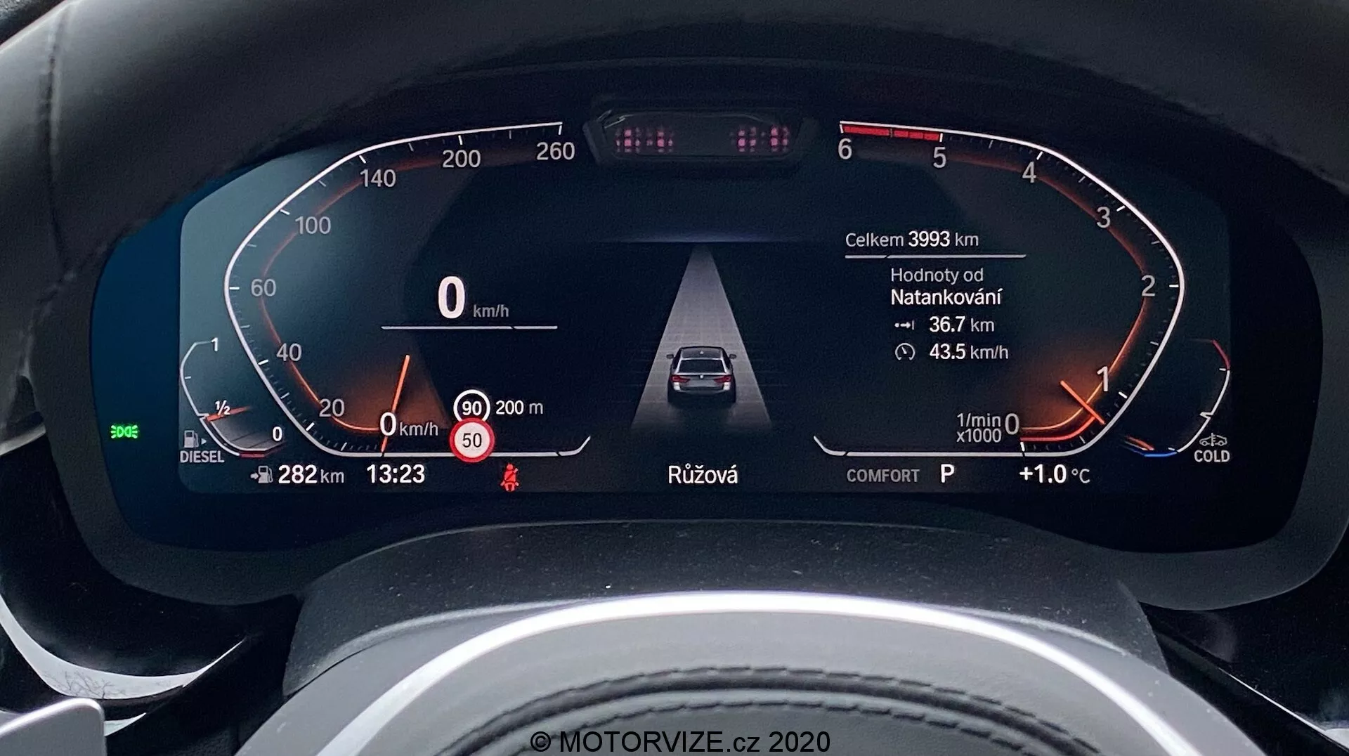 Zbliżenie na cyfrową deskę rozdzielczą BMW Serii 5 2020 (BMW 520i Touring Steptronic) z perspektywy kierowcy. Cyfrowy panel instrumentów wyświetla kluczowe informacje, takie jak prędkość pojazdu (0 km/h), wskaźnik paliwa oznaczony dla oleju napędowego, ostrzeżenia o otwartych drzwiach i przypomnienie o pasach bezpieczeństwa, zaawansowane systemy wspomagania kierowcy, takie jak adaptacyjny tempomat, obrotomierz w stanie jałowym z wskaźnikiem zimnego silnika oraz dodatkowe dane, takie jak przebieg (39933 km), temperatura zewnętrzna (+1.0°C), czas (13:23), wybór biegu (P dla parkowania) i tryb jazdy (komfortowy). Projekt jest elegancki i nowoczesny, co wskazuje na samochód wysokiej klasy.