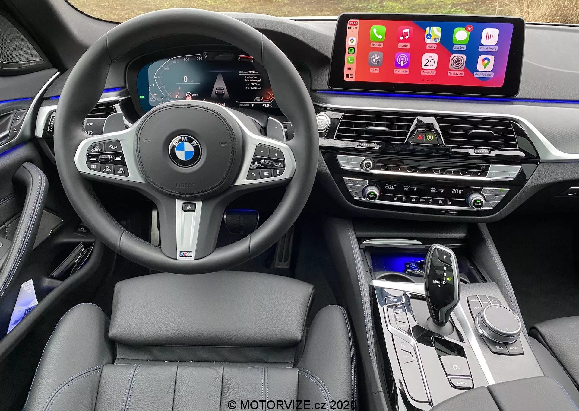 Widok wnętrza z perspektywy fotela kierowcy BMW 520i Touring Steptronic z 2020 roku, z wielofunkcyjną kierownicą obszytą skórą z logo BMW i przyciskami sterowania tempomatem, audio i telefonem. Cyfrowy zestaw wskaźników pokazuje prędkościomierz, obrotomierz i mapę nawigacyjną. Konsola środkowa wyposażona jest w duży dotykowy system infotainment, wyświetlający aplikacje mobilne, takie jak Apple CarPlay. Poniżej znajdują się otwory wentylacyjne, panel sterowania klimatyzacją, sterowanie audio, elegancka dźwignia zmiany biegów i przyciski funkcyjne. Wnętrze pokazuje skórzane fotele, oświetlenie otoczenia i wysokiej jakości materiały z metalicznymi akcentami.