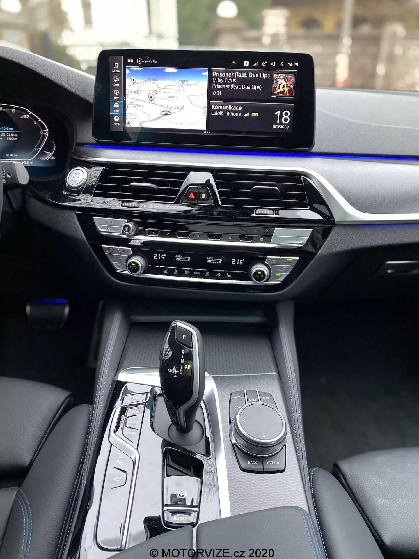 Widok wnętrza BMW Serii 5 2020 (BMW 520i Touring Steptronic), ukazujący konsolę centralną i deskę rozdzielczą z perspektywy przedniego pasażera. Konsola centralna zawiera nowoczesny selektor automatycznej skrzyni biegów w trybie 'park', z różnymi elementami sterującymi, w tym obrotowym pokrętłem do systemu informacyjno-rozrywkowego oraz przyciskami do trybów jazdy lub ustawień pojazdu. Środkowa część deski rozdzielczej obejmuje sterowanie klimatyzacją z intuicyjnymi pokrętłami i przyciskami, powyżej których znajduje się slot na CD. Górną część deski rozdzielczej zajmuje szeroki ekran dotykowy systemu informacyjno-rozrywkowego z mapą nawigacji i informacjami o ograniczeniach prędkości. Częściowo widoczny jest cyfrowy zestaw wskaźników z prędkościomierzem. Materiały wykończeniowe wnętrza podkreślają luksusowy charakter, kontrastując ciemną skórę z niebieską listwą akcentującą na całej szerokości deski rozdzielczej.
