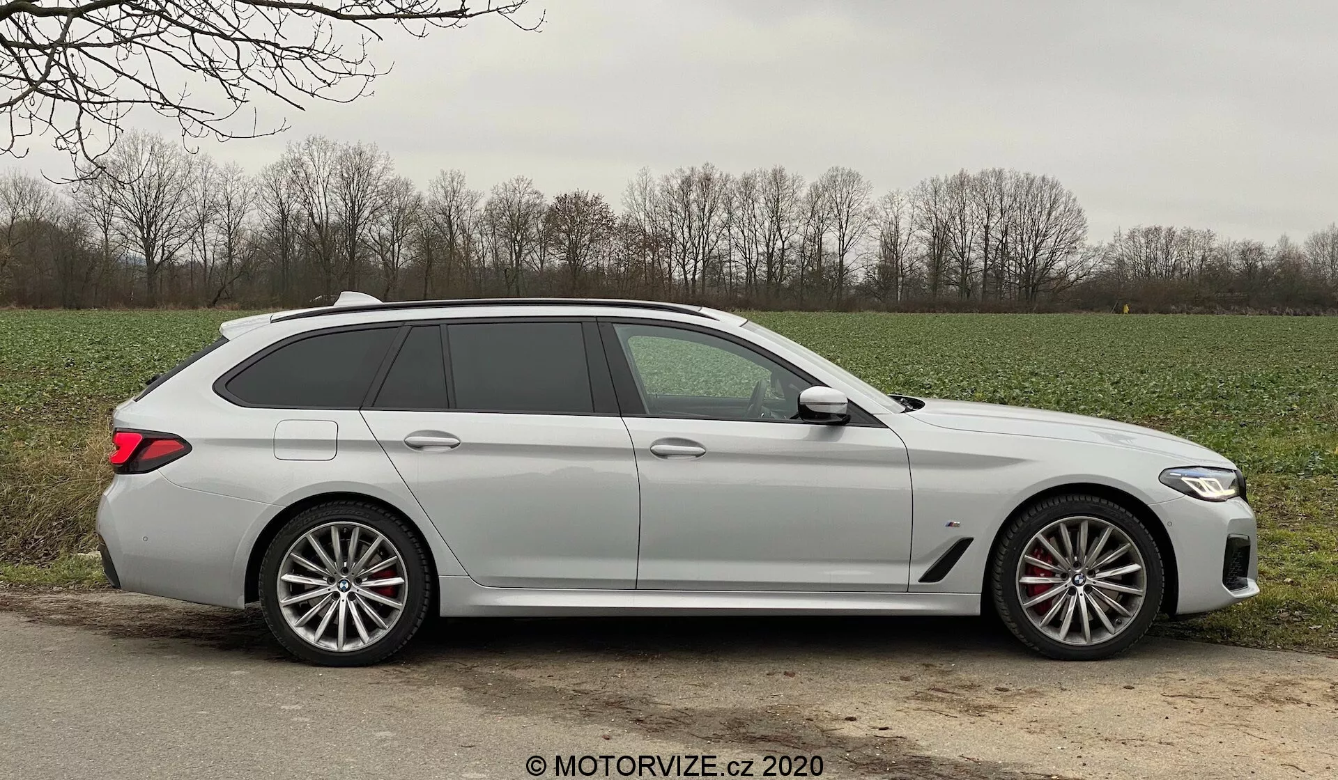 Profil boczny nowoczesnego kombi BMW 520i Touring Steptronic uchwycony w świetle dziennym, ukazujący eleganckie linie projektowe, pięć drzwi w tym tylna klapa, długi dach ze spojlerem, duże wieloramienne felgi ze stopów metali lekkich z widocznymi zaciskami hamulców, klamki w kolorze nadwozia, zintegrowane kierunkowskazy w lusterkach bocznych, lekko przyciemniane szyby i matowe lub niemetaliczne wykończenie lakieru. Samochód jest ustawiony na szutrowym lub ziemnym poboczu obok asfaltowej drogi w wiejskim otoczeniu, z trawą i drzewami w tle, przy sprzyjających warunkach pogodowych.