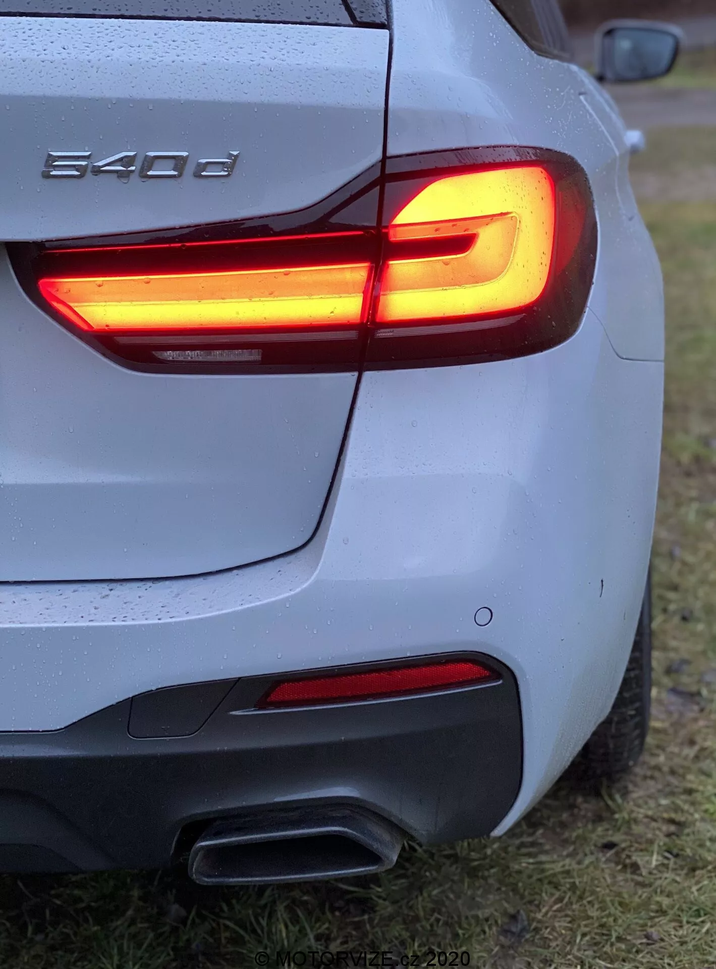 Zbliżenie na tylny trzykwaratałowy kąt BMW 540i, prezentujące tylną lampę LED w kształcie litery L, podwójne rury wydechowe, srebrne/szare nadwozie z kroplami wody, rzeźbiony tylny zderzak, częściowe logo BMW, zaparkowane na trawiastej powierzchni.