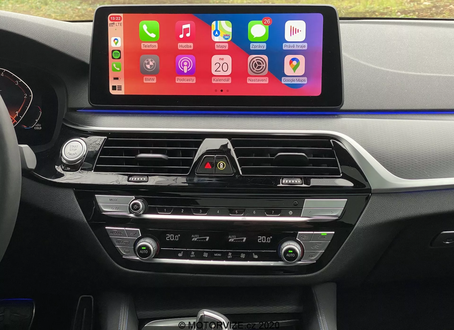 Zdjęcie wnętrza BMW 5er-Reihe 2020 (BMW 520i Touring Steptronic) z perspektywy fotela kierowcy, skupiające się na centralnej desce rozdzielczej. Najbardziej widocznym elementem jest duży, poziomo ustawiony ekran dotykowy systemu infotainment z kolorowym interfejsem wspierającym integrację smartfonów, takich jak Apple CarPlay i Android Auto. Poniżej ekranu widać otwory wentylacyjne z chromowanymi akcentami oraz przycisk świateł awaryjnych. Niżej znajdują się sterowniki klimatyzacji dwustrefowej z cyfrowymi wyświetlaczami ustawień temperatury, przyciskami i pokrętłami do regulacji temperatury powietrza, prędkości wentylatora i rozprowadzania powietrza. Deska rozdzielcza i konsola centralna wykonane są z miękkiego plastiku oraz metalicznych akcentów, co podnosi atrakcyjność estetyczną wnętrza.