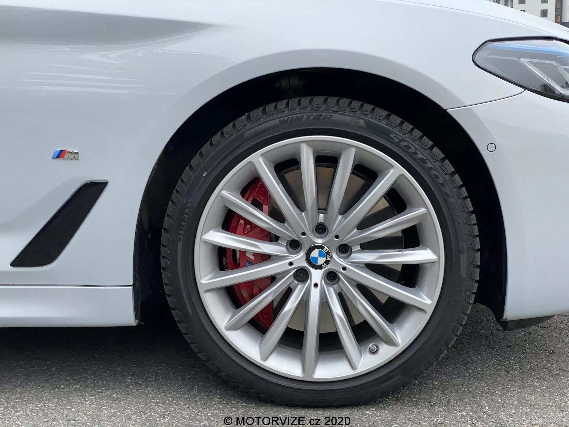 Zbliżenie na przednią część koła BMW z Serii 5 z 2020 roku, konkretnie modelu 520i Touring Steptronic. Zdjęcie z niskiego kąta podkreśla skomplikowany wieloszprychowy design felgi z srebrnym wykończeniem, uzupełniony czerwoną zaciskiem hamulcowym i wentylowanym rotorem. Znak BMW i odznaka 'M' na błotniku wskazują, że jest to pojazd zorientowany na osiągi z serii M BMW. Obraz uchwytuje kluczowe szczegóły odpowiednie do przedstawienia wysokowydajnych systemów hamulcowych, designu luksusowych pojazdów i technologii opon.
