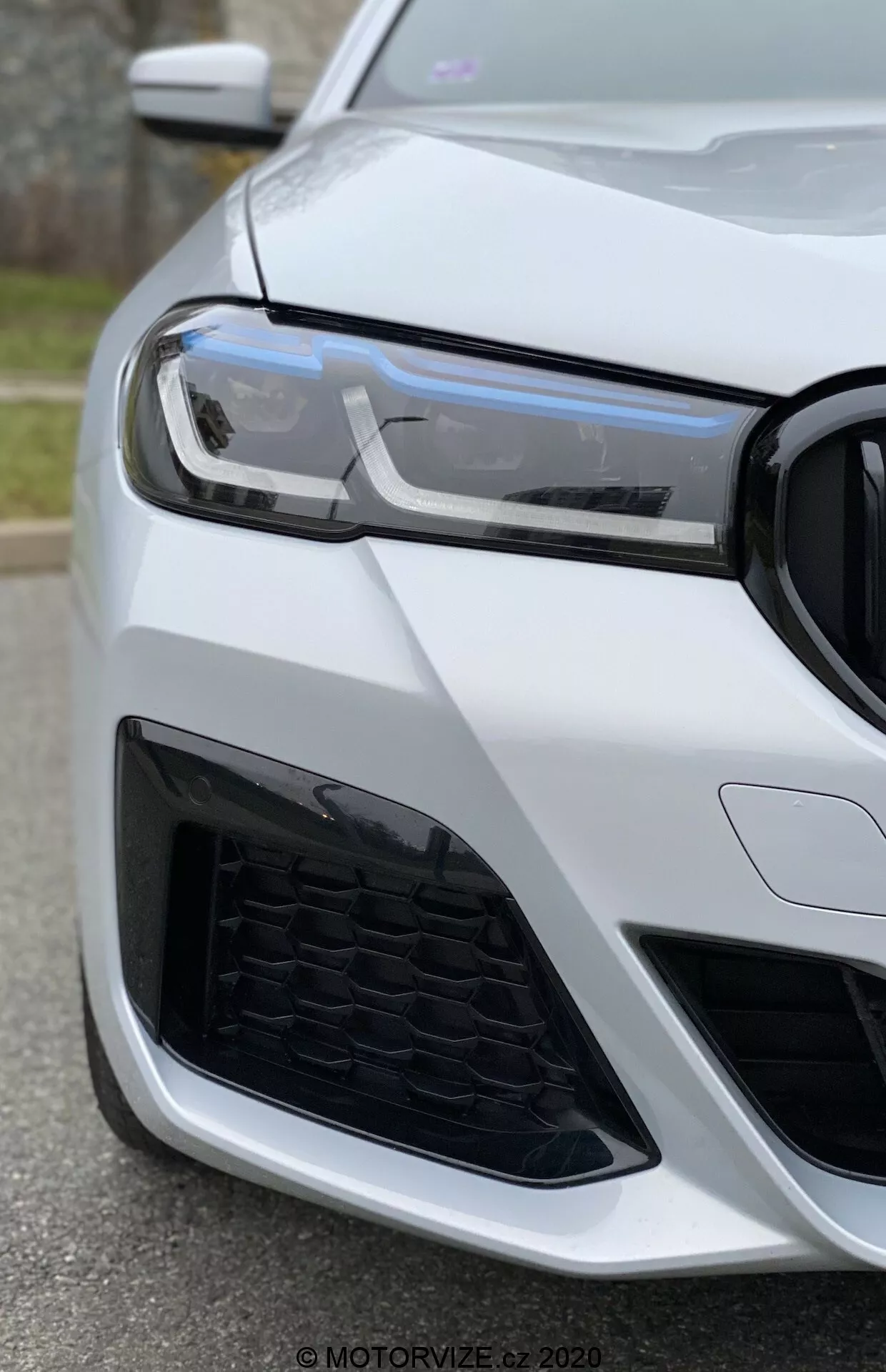 Zbliżenie lewego przedniego rogu BMW 520i Touring Steptronic (2020) pod kątem z dołu, skupiając się na reflektorze, grillu i zderzaku. Nowoczesne światła dzienne LED, wzór plastra miodu na grillu i agresywny design wlotu powietrza sugerują sportowy i zaawansowany pojazd. Samochód jest pomalowany na błyszczący biały kolor, podkreślając luksusowy i dobrze utrzymany wygląd.