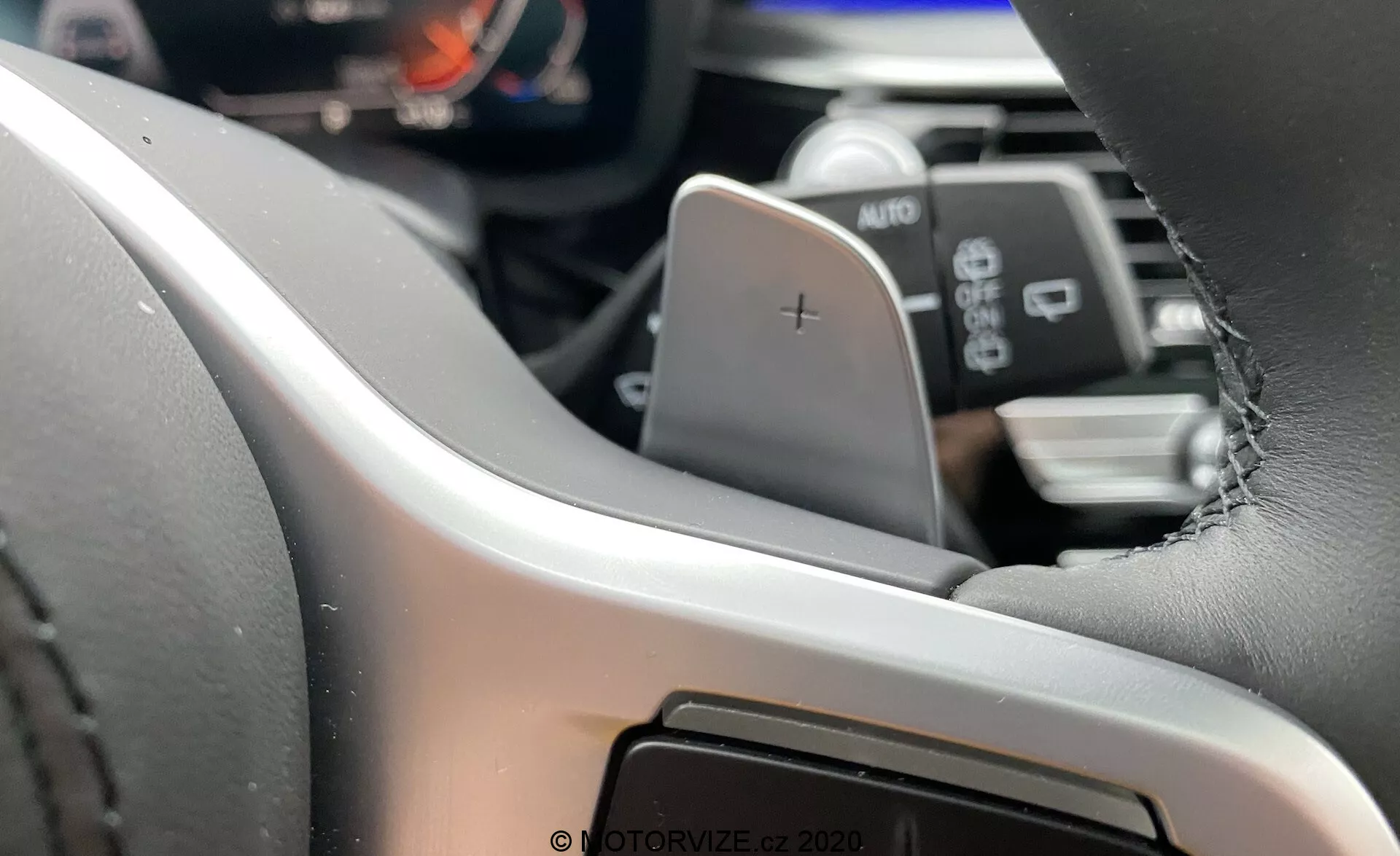 Widok wnętrza BMW Serii 5 2020 (BMW 520i Touring Steptronic) skupiający się na lewej stronie kierownicy z perspektywy kierowcy⁠—nowoczesna kierownica obszyta skórą z widocznymi szwami, wyposażona w łopatkę zmiany biegów oznaczoną '+'. Cyfrowy wyświetlacz lub panel przyrządów jest częściowo widoczny, podkreślając zaawansowane systemy informacyjne dla kierowców. Poniżej łopatki znajduje się dźwignia oznaczona AUTO, która prawdopodobnie kontroluje światła lub wycieraczki. W rozmytym tle widać fragment systemu multimedialnego lub konsoli centralnej, sugerując interfejs dotykowy.