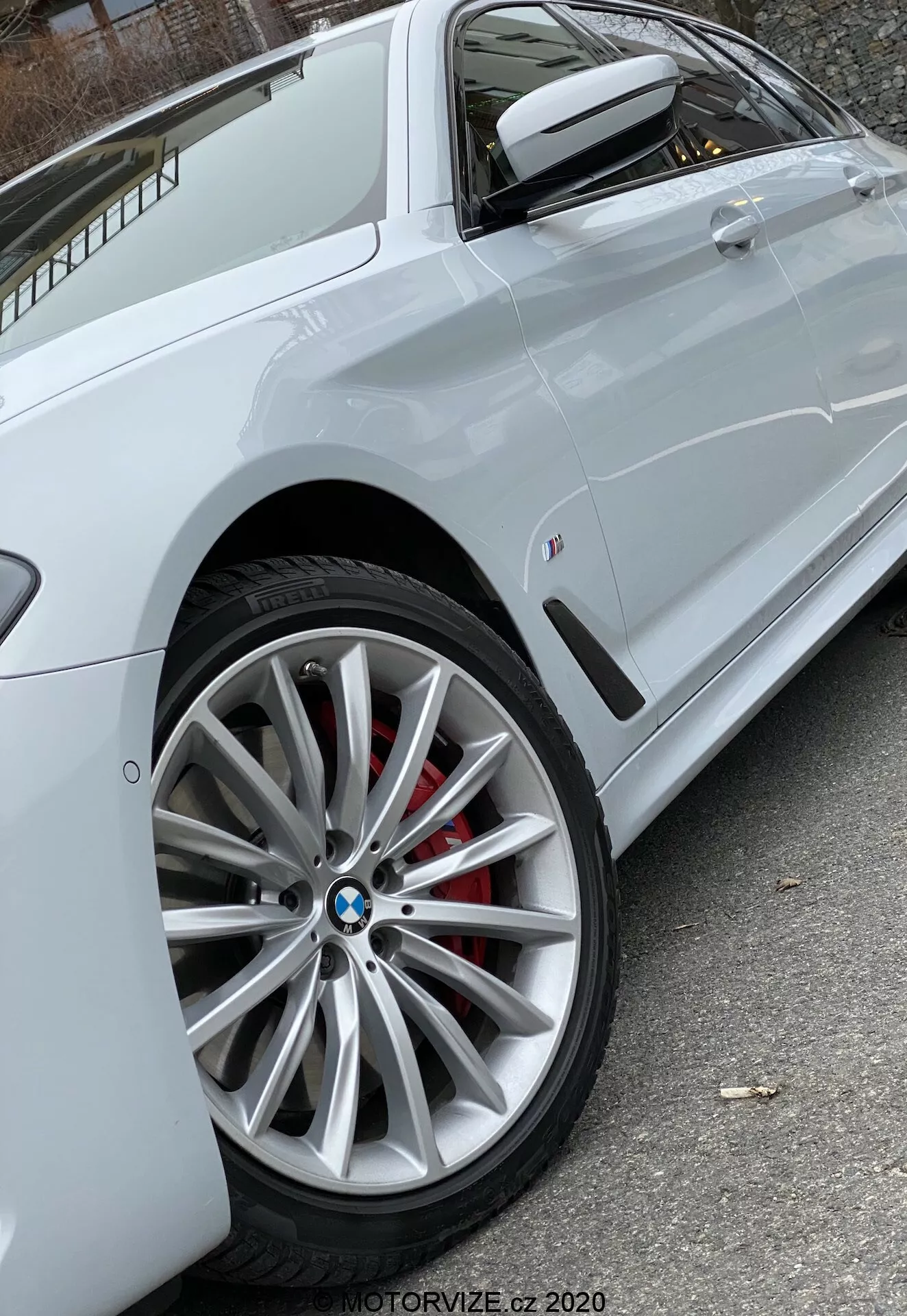 Bliskie, skośne ujęcie boczne białego BMW 520i Touring Steptronic (2020) skupiające się na nowoczesnym przednim kole wieloramiennym z widocznym logo BMW i czerwonym zaciskiem hamulcowym, częściowo ukazujące bok z wyrazistą linią charakteru biegnącą przez klamki drzwi, lusterkiem bocznym w kolorze nadwozia z wbudowanym kierunkowskazem, oraz malutką plakietką na przednim panelu bocznym. Naturalne oświetlenie zapewnia wyraźną widoczność cech pojazdu. Zdjęcie autorstwa MOTORVIZE.cz 2020.