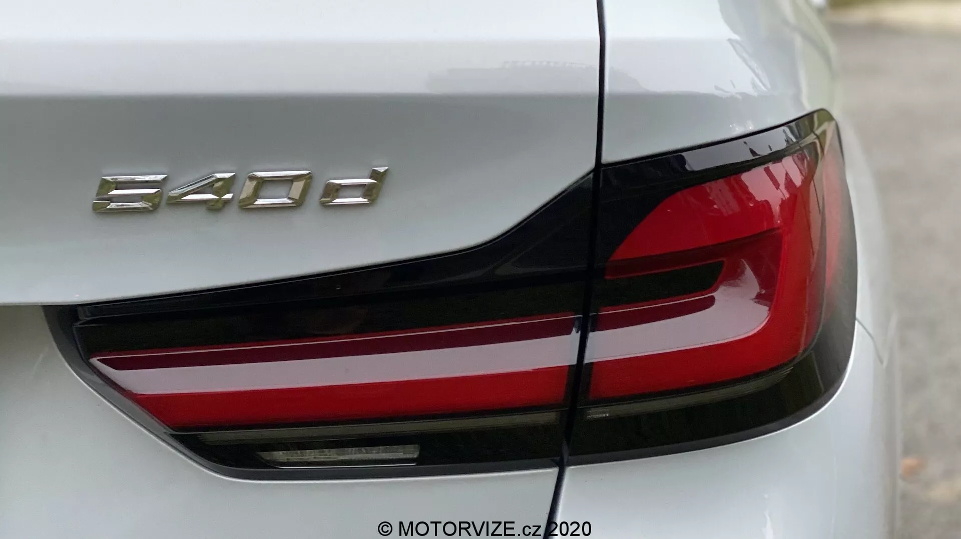 Zbliżenie tylnej części BMW 520i Touring Steptronic, koncentrując się na lewym tylnym świetle i części bagażnika. Kąt jest lekko ukośny od lewego tylnego rogu, podkreślający nowoczesne tylne światło LED o ostrym, kanciastym designie i wyraźnej czerwonej sekcji. Pokrywa bagażnika pokazuje oznaczenie modelu '840d.' Karoseria samochodu jest błyszcząca, prawdopodobnie biała lub jasnosrebrna, podkreślając luksus i precyzyjne wykonanie.