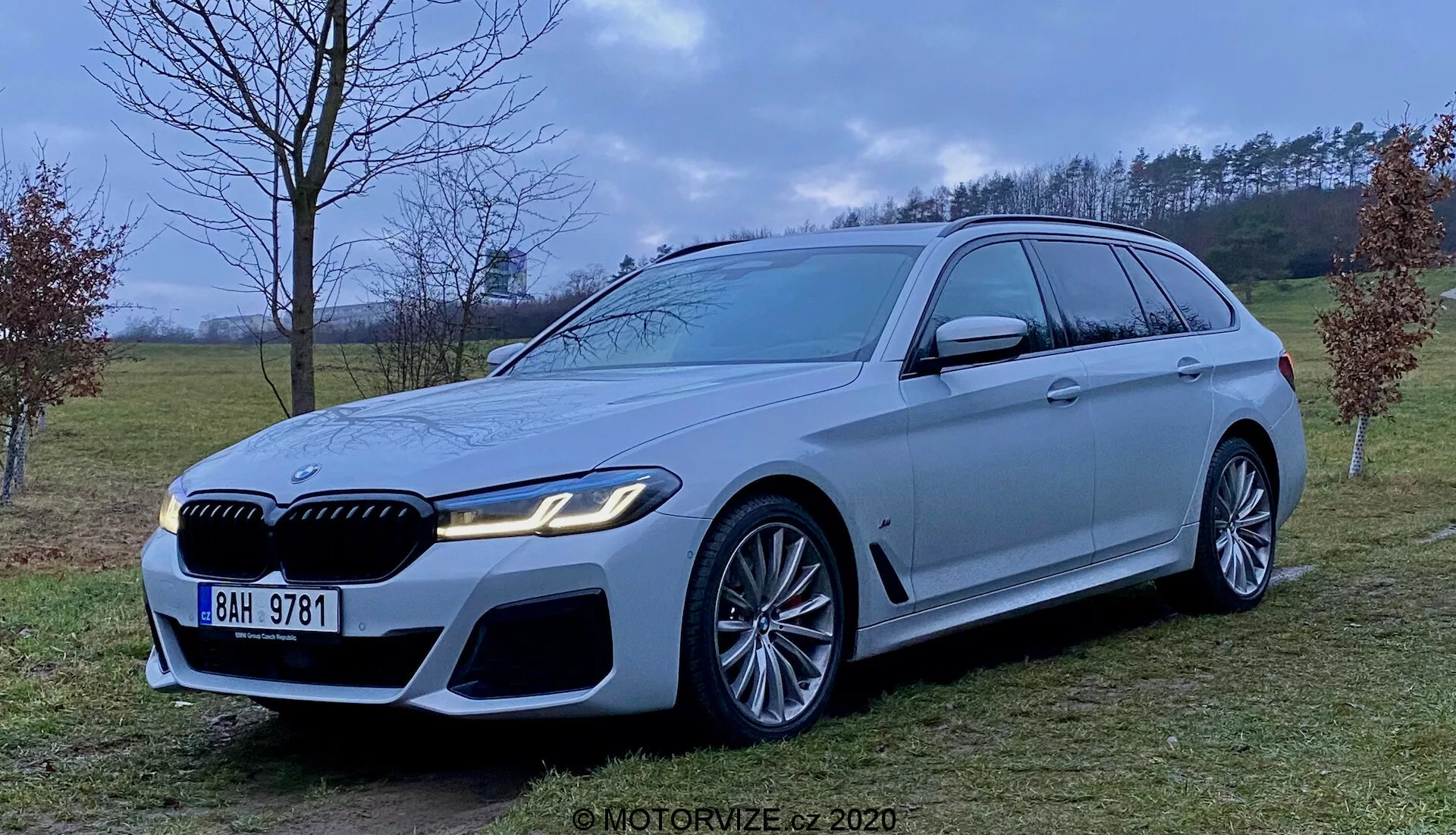 Obraz luksusowego kombi BMW serii 5 Touring 2020 (BMW 520i Touring Steptronic) w jasnosrebrnym lub szarym kolorze, ukazującego agresywną i sportową przednią część z dużymi wlotami powietrza, wyraźnym grillem i eleganckimi reflektorami LED. Pojazd ma duże felgi aluminiowe z wieloramiennym wzorem i wyraźnymi zaciskami hamulcowymi, długą linię dachu ze spoilerem, chromowane listwy okien oraz silną linię barkową. Zdjęcie wykonano z wysokości w połowie pojazdu w półwiejskim otoczeniu z rozproszonym oświetleniem.