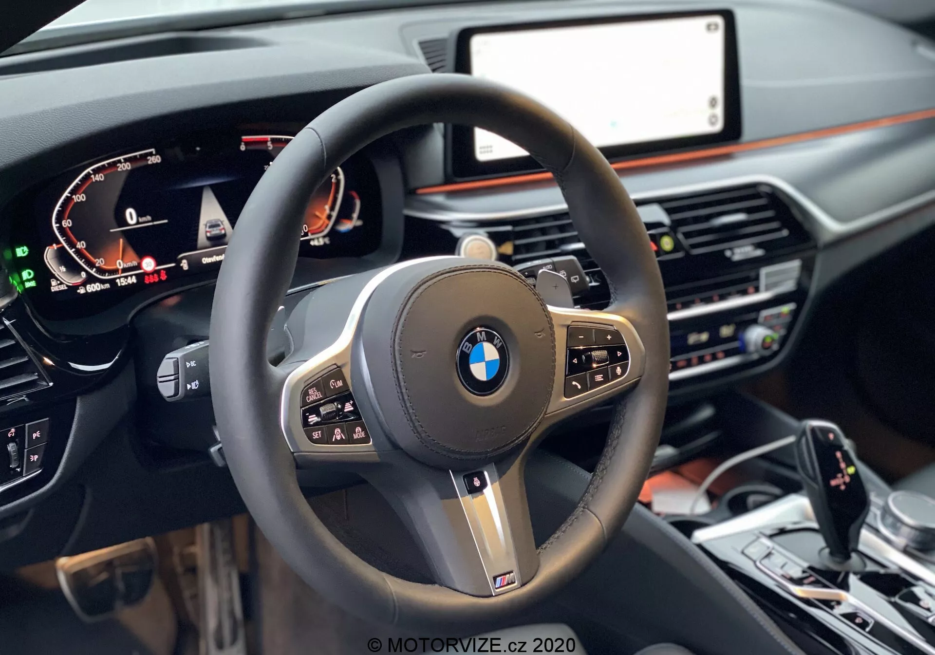 Widok z perspektywy pierwszej osoby z siedzenia kierowcy w BMW 520i Touring Steptronic 2020, skupiający się na nowoczesnej kierownicy trzema ramionami z grubą, skórzaną obręczą, wielofunkcyjnymi przyciskami i wyraźnym logo BMW pośrodku, otoczonym błyszczącymi czarnymi i metalicznymi akcentami. Cyfrowa tablica przyrządów po lewej stronie wyświetla prędkość, obroty i informacje dla kierowcy, podczas gdy po prawej, częściowo widać centralny ekran infotainment. Deska rozdzielcza o ciemnych odcieniach z dodatkiem drewna zawiera poniżej ekranu kontrolki klimatyzacji i systemu multimedialnego. Na dole obrazu widać konsolę centralną z wybierakiem biegów, kontrolerem iDrive i przyciskami dynamicznego kontrolowania jazdy.