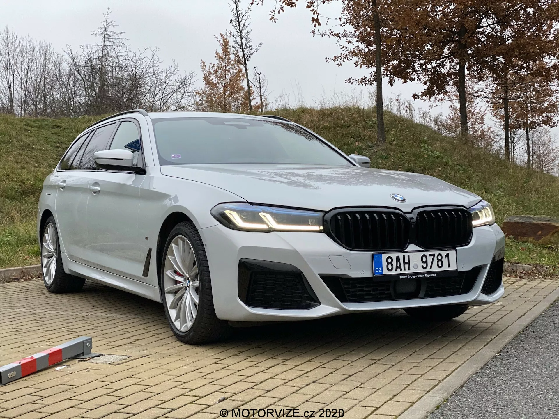 Obraz przedstawia BMW Serii 5 z 2020 roku (520i Touring Steptronic), nowoczesny luksusowy sedan, zaparkowany na lekkim wzniesieniu. Zdjęcie wykonano z niskiego i nieco bocznego kąta, ukazując przedni kwartał oraz profil pojazdu. Zauważalne detale to wyrazista atrapa chłodnicy w kształcie nerek, złożone wloty powietrza w przednim zderzaku, eleganckie, kątowe reflektory LED, duże, wieloramienne felgi aluminiowe, długa, płynna sylwetka nadwozia z metalicznym, srebrnym wykończeniem, chromowane akcenty, zintegrowane kierunkowskazy w lusterkach bocznych oraz potencjalne zaawansowane technologie, takie jak asystenci jazdy autonomicznej i asysta parkowania. Na przednim panelu bocznym widoczny jest emblemat wskazujący na konkretną wariantę modelu.