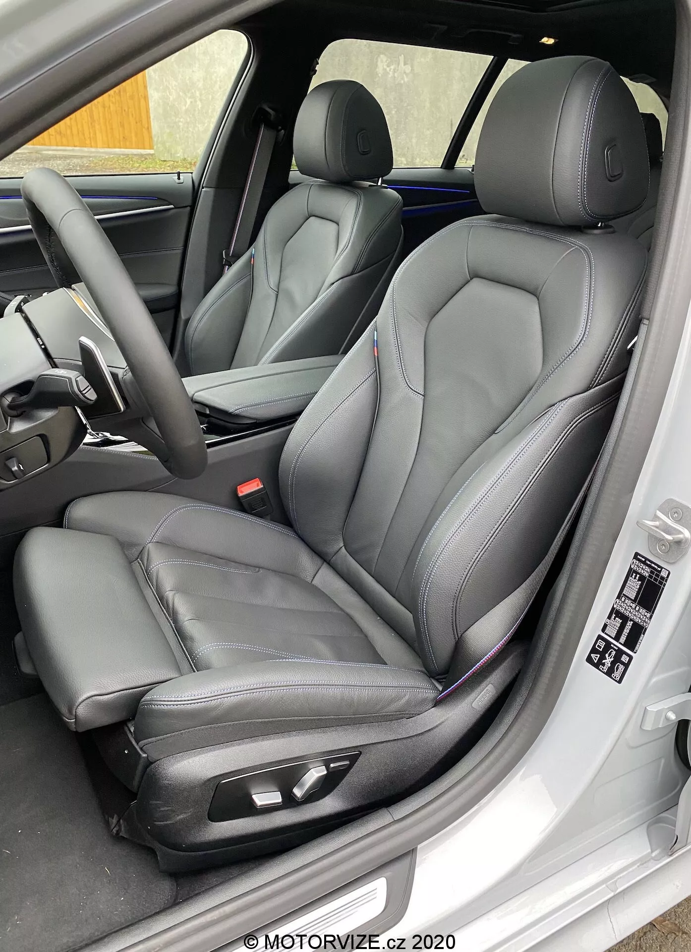 Zbliżenie ukośne na fotel kierowcy BMW 520i Touring Steptronic 2020, prezentujące nowoczesne przednie fotele typu kubełkowego z czarnej skóry z kontrastującymi szwami, wyposażone w elektroniczne sterowanie dla zwiększenia komfortu. Obraz ukazuje również wewnętrzny panel drzwi kierowcy z elektrycznymi sterownikami szyb i lusterek, ustawieniami pamięci fotela oraz dodatkowymi cechami deski rozdzielczej i kierownicy, wskazującymi na luksus i zaawansowaną technologię.