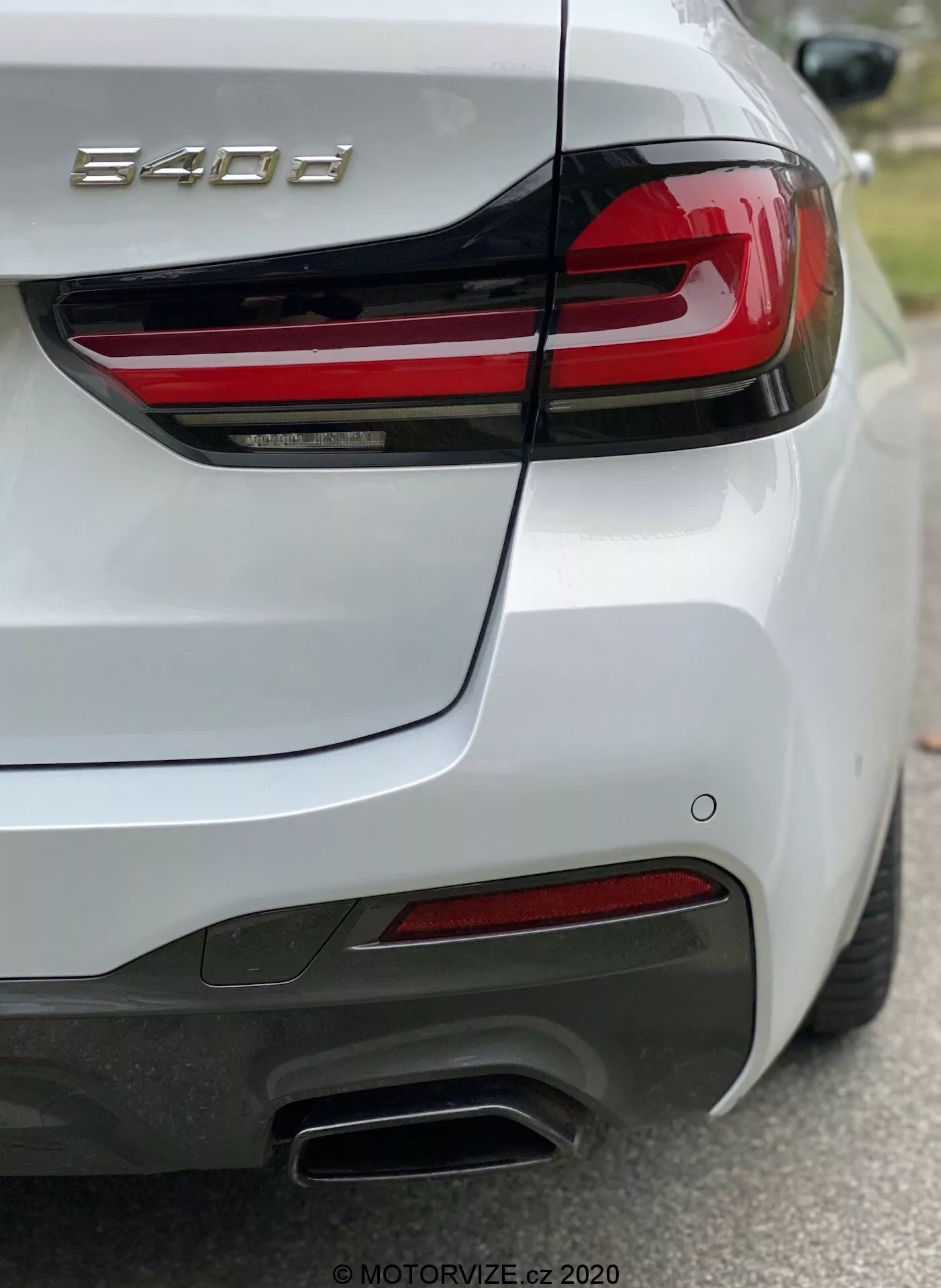 Tył samochodu BMW serii 8 (840i), uchwycony z tylnego prawego kwartału, podkreślający prawy tylny światła z poziomymi czerwonymi paskami LED, rzeźbioną pokrywę bagażnika z wbudowanym spojlerem, tylny zderzak z czujnikami, podwójne metaliczne końcówki wydechu po prawej stronie, czerwone odblaski i białą błyszczącą farbę. Zdjęcie skupia się na oznaczeniu modelu '840i', pokazując szczegóły projektowe i cechy technologiczne, takie jak oświetlenie i systemy wspomagające kierowcę.