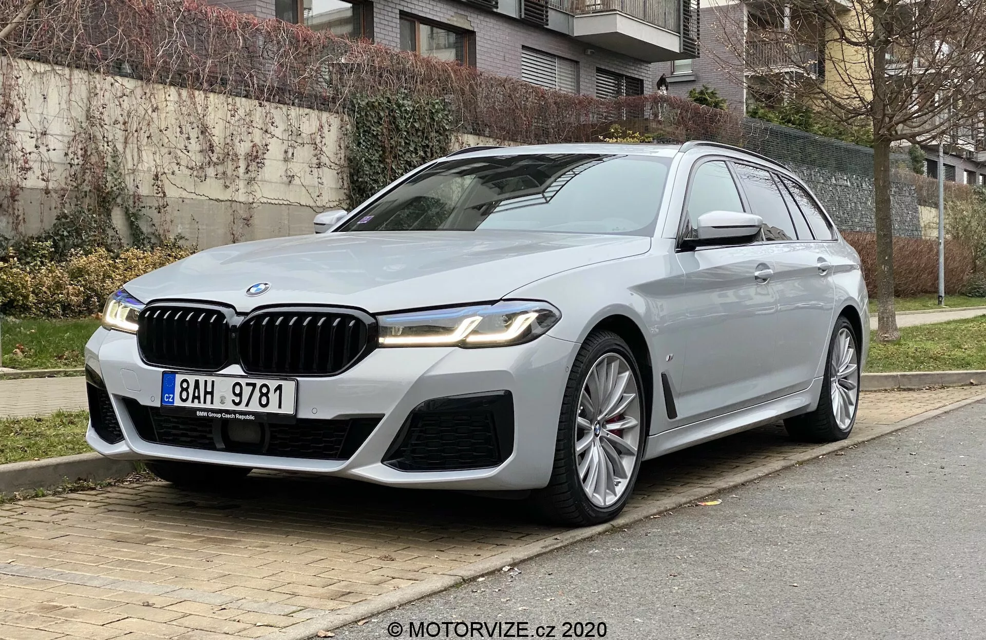 Luksusowe BMW 520i Touring zaparkowane na miejskiej ulicy, widziane z niskiego przedniego trójkątnego ujęcia, podkreślającego przód i bok kierowcy. Samochód cechuje się prominantną kratką nerkową z chromowanymi detalami, eleganckimi reflektorami LED, sportowym przednim zderzakiem i wyraźnymi zagięciami na masce. Wieloramienne felgi aluminiowe, lusterka boczne z kierunkowskazami i kamerą, chromowane ramy okien oraz szyberdach dodają luksusowego wyglądu. Ustawiony po prawej stronie z trawą, krzewami i budynkami mieszkalnymi w tle sugerują zaawansowaną technologię i ekskluzywny design.