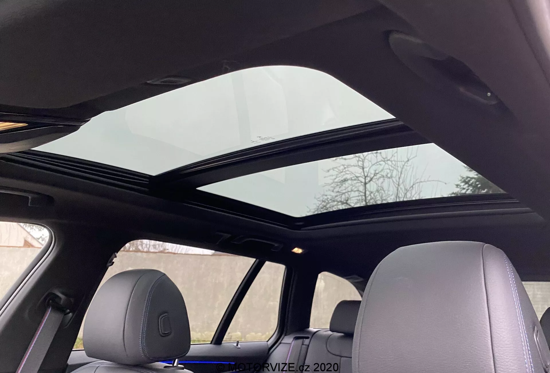Widok wnętrza BMW Serii 5 2020 520i Touring z przekładnią Steptronic z perspektywy pasażera tylnego siedzenia, spoglądającego w górę, ukazujący panoramiczny szyberdach rozciągający się nad przednimi i tylnymi siedzeniami. Przedni panel szyberdachu jest lekko otwarty, poprawiając wentylację. Podsufitka ma jasny kolor, komponując się z wnętrzem pojazdu, z zagłówkami dopasowanymi do materiału wnętrza, podkreślając zaawansowany komfort i nowoczesne udogodnienia.