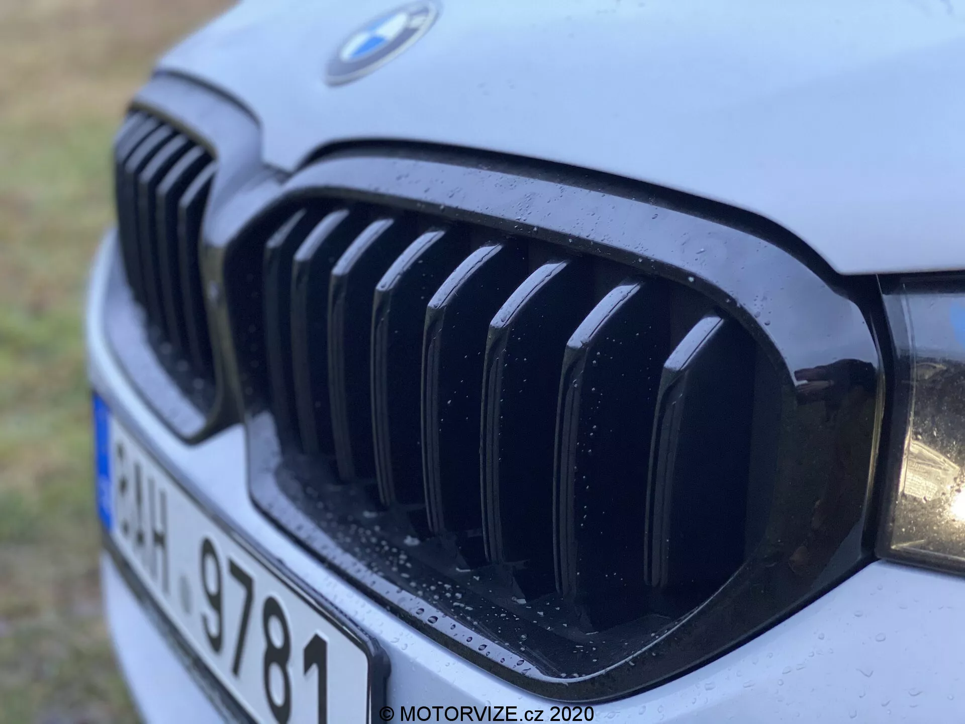 Zbliżenie przedniego grilla BMW 520i Touring Steptronic z roku 2020, ukazujące charakterystyczny grill nerkowy i emblemat BMW z pionowymi czarnymi listwami, częściowo widoczny reflektor i krople wody sugerujące niedawny deszcz lub mycie, uchwycone pod niskim kątem z lewej przedniej strony.